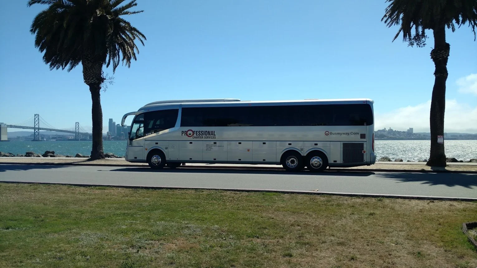 A white bus labeled 'Professional Charter Services' parked on a waterfront road, with a city skyline and San Francisco Bay Bridge in the background, under a partly cloudy sky.