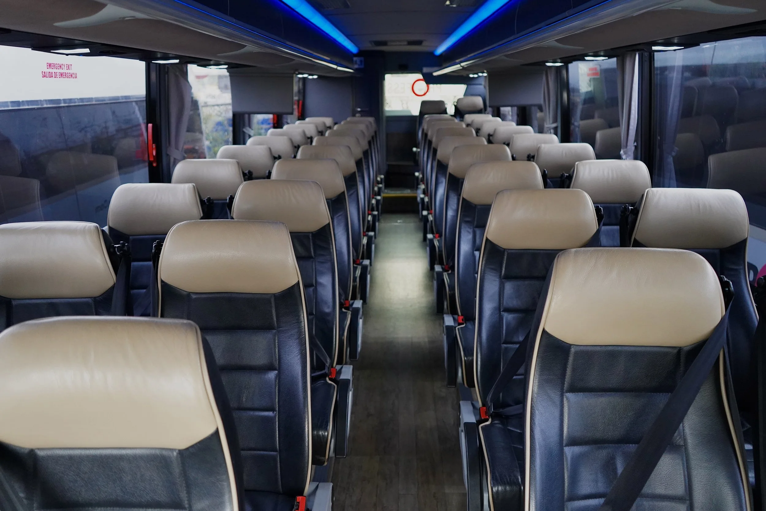 Empty bus interior with rows of beige and black leather seats, overhead lighting, windows with curtains, and emergency exit signs.