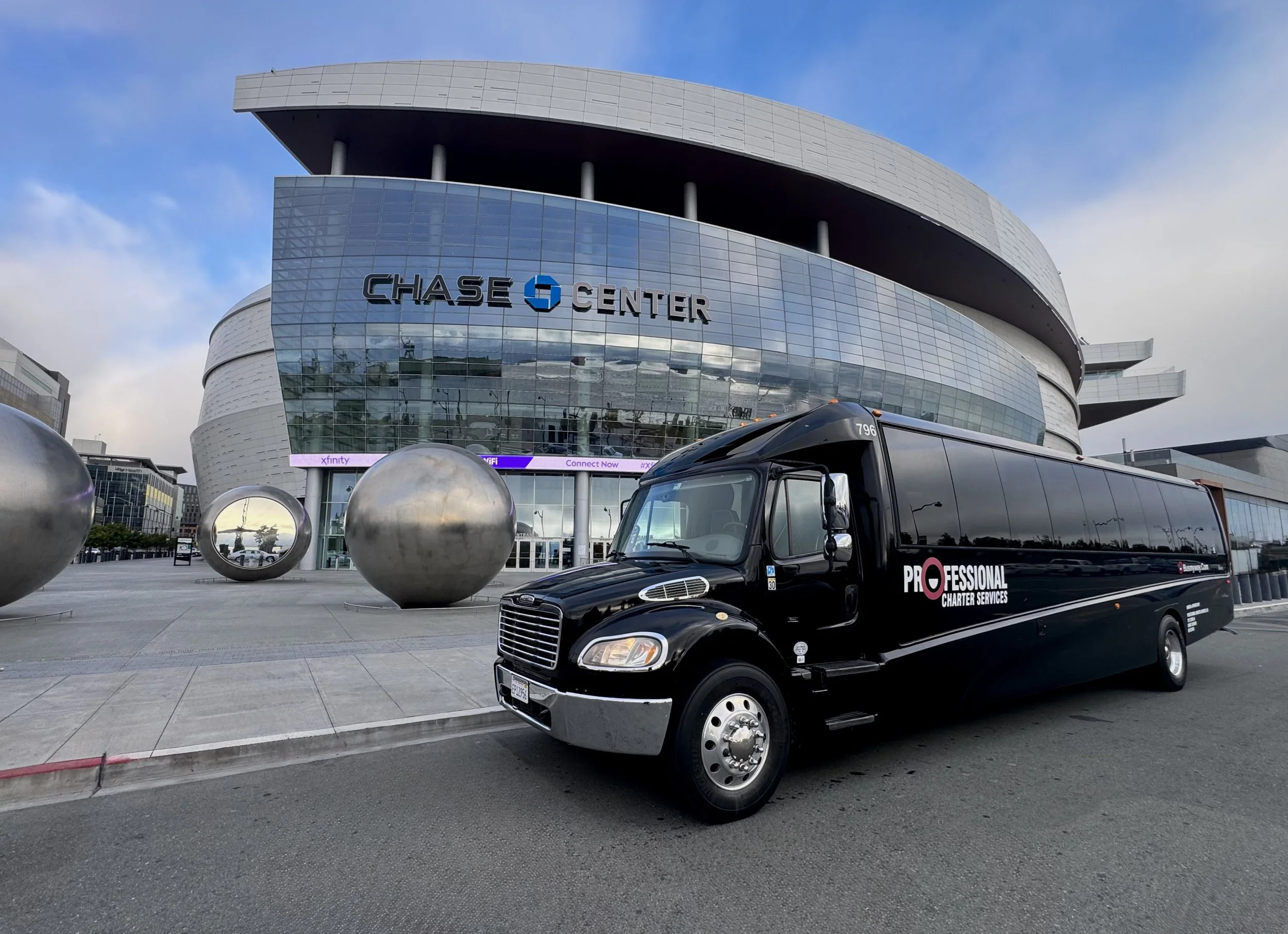 Black professional charter services bus parked in front of Chase Center with large metallic spheres on the sidewalk and the modern glass building in the background.
