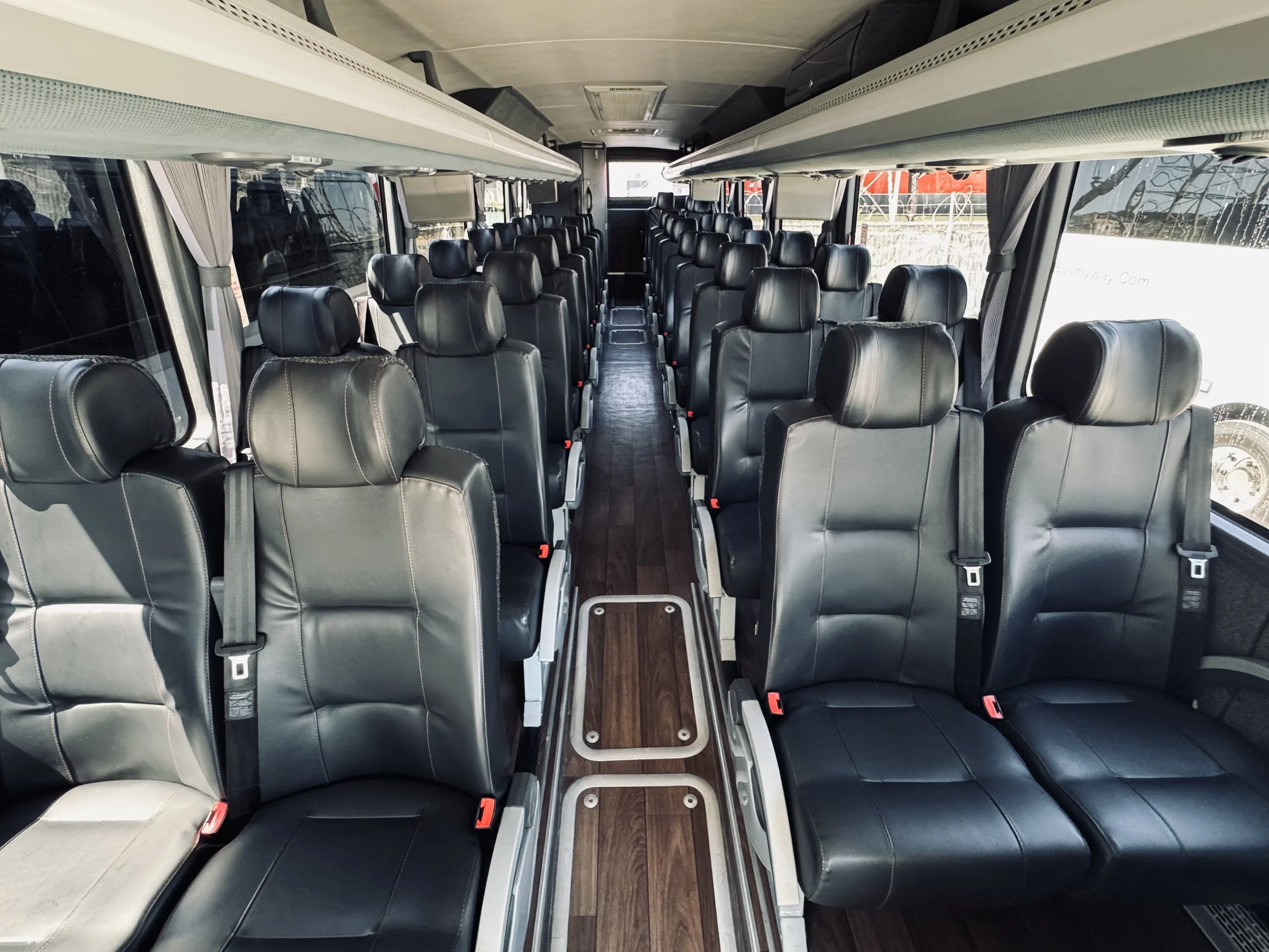 Interior view of a bus with black leather seats, an aisle in the middle, large windows on both sides, and overhead storage compartments.