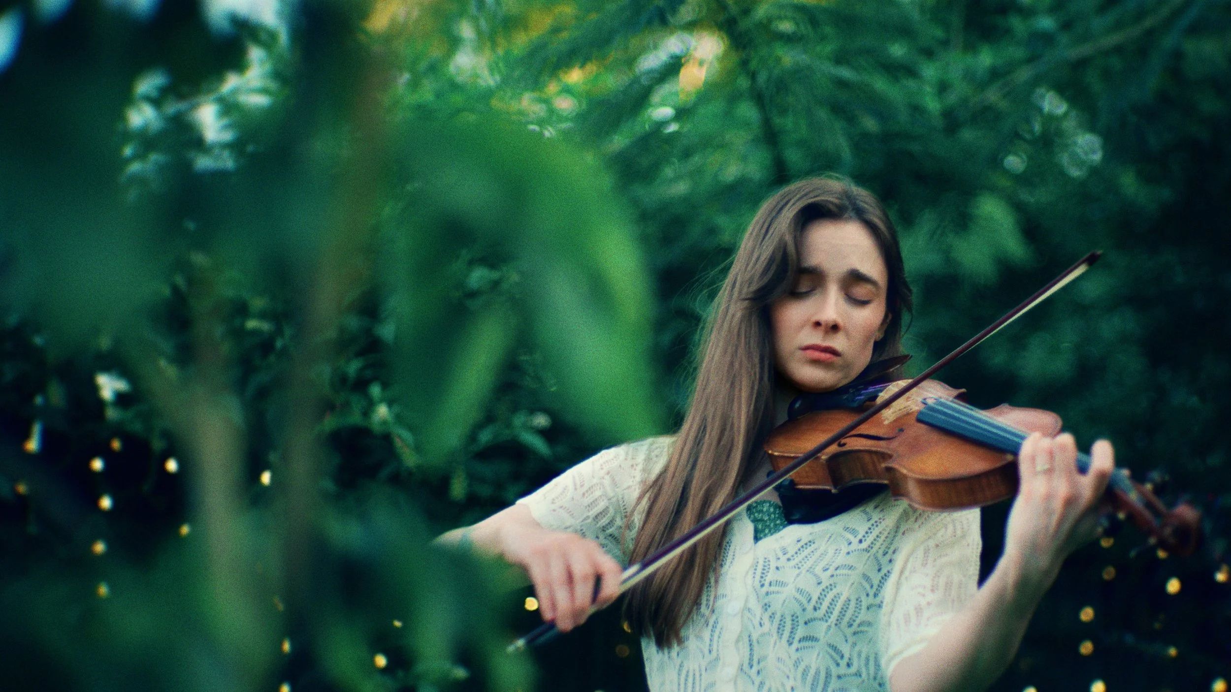A woman with long brown hair wearing a white lace top is playing the violin outdoors among green trees.