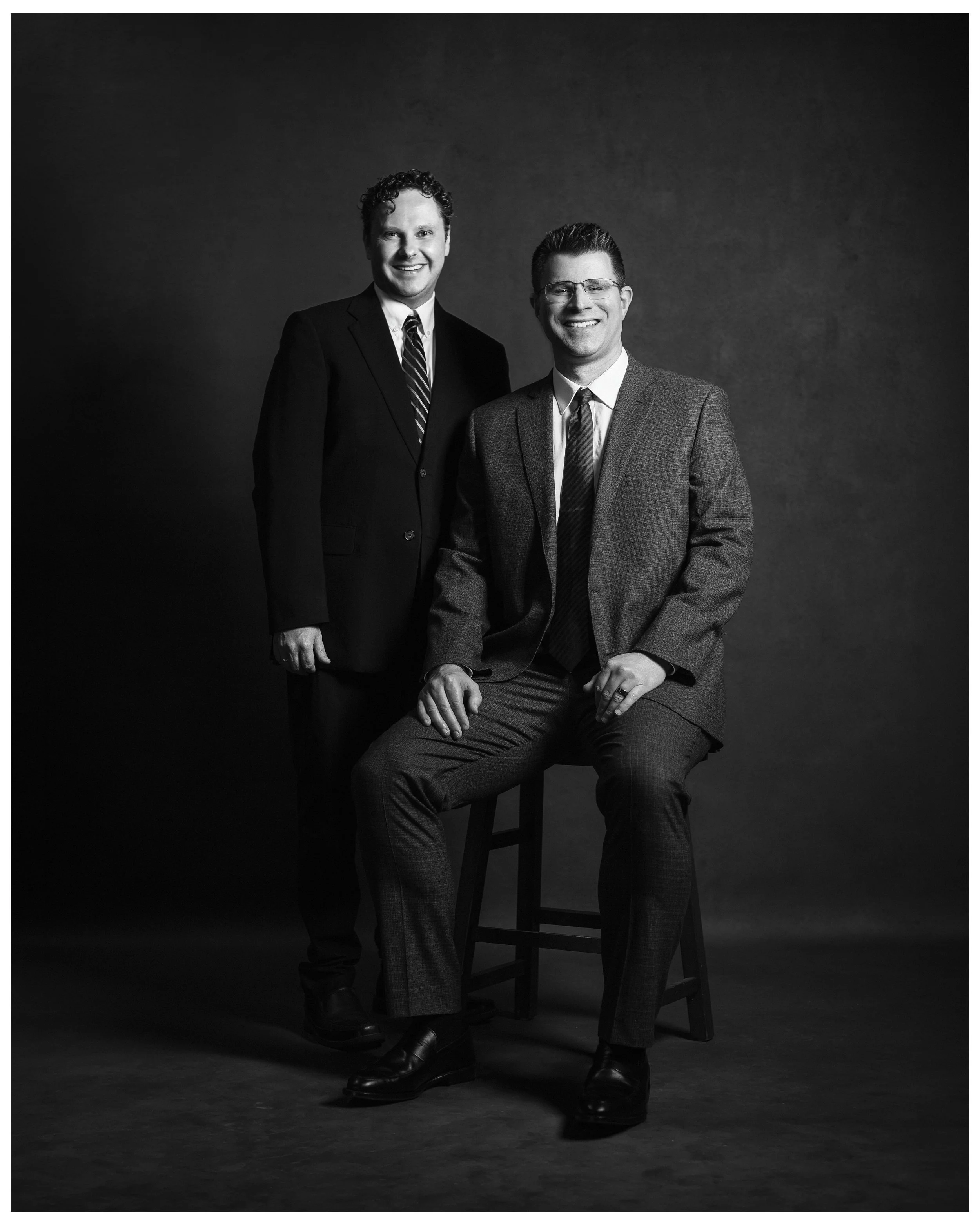 Black and white photo of two men in suits, one sitting on a wooden stool and the other standing behind, both smiling, against a dark plain background.