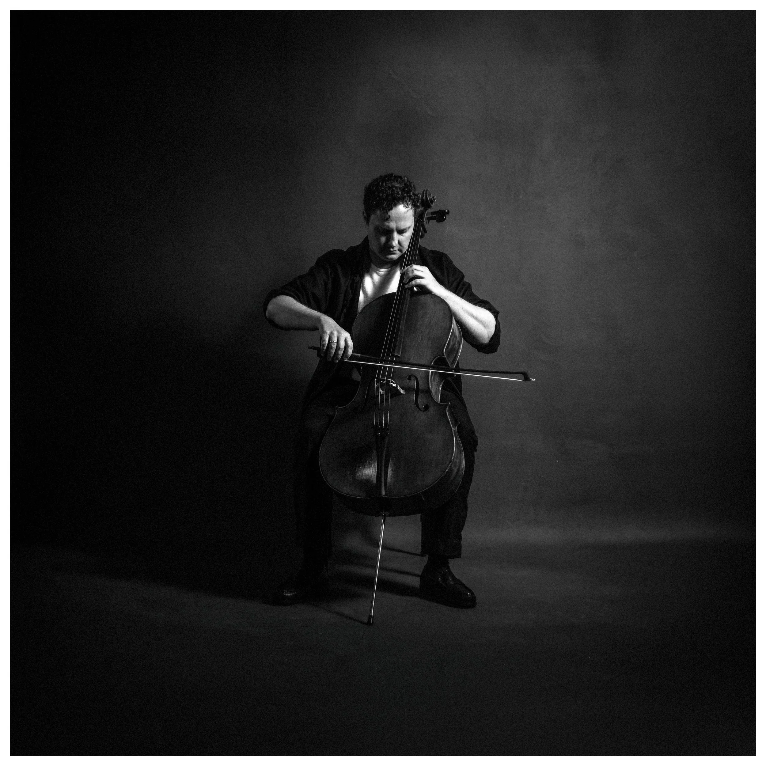A person playing the cello, standing against a dark background with minimal lighting.