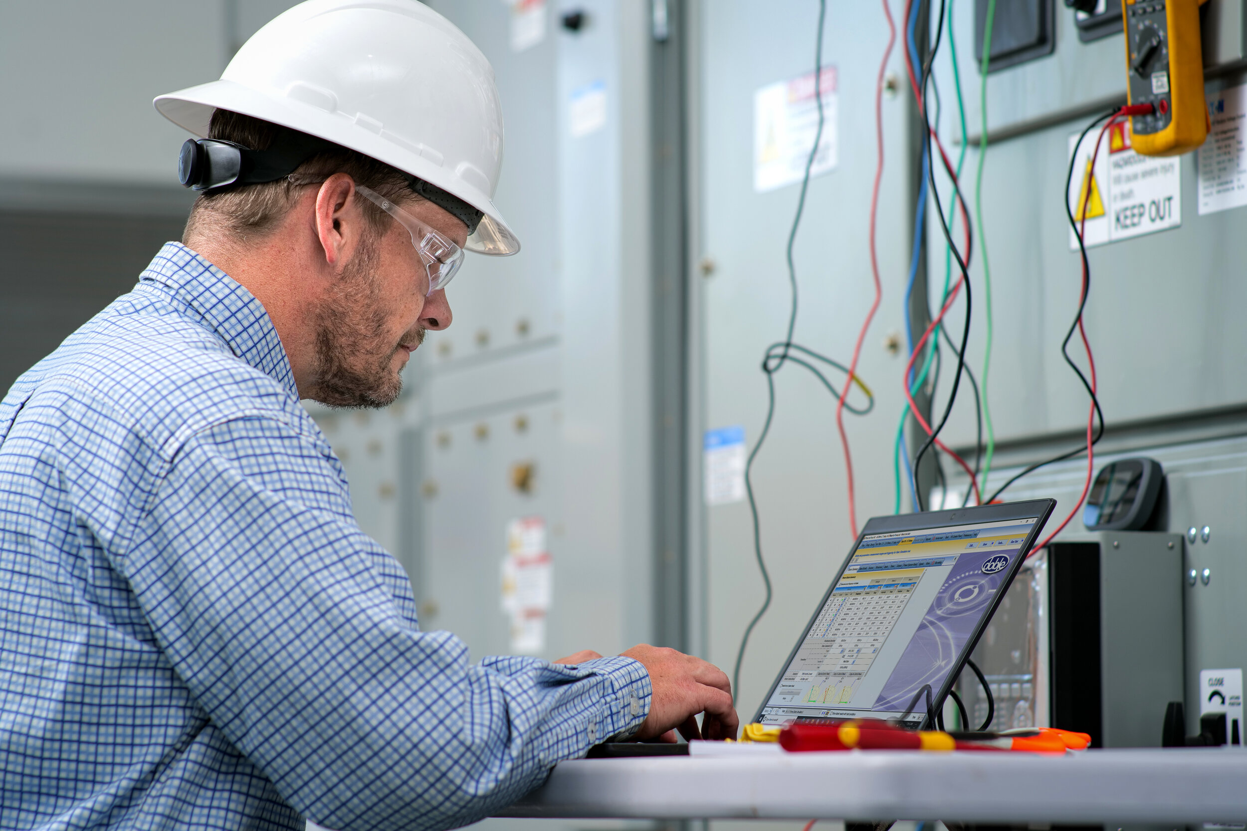 Male engineer wearing hard hat and safety glasses using a laptop near electrical panels, with wires and tools in the background.