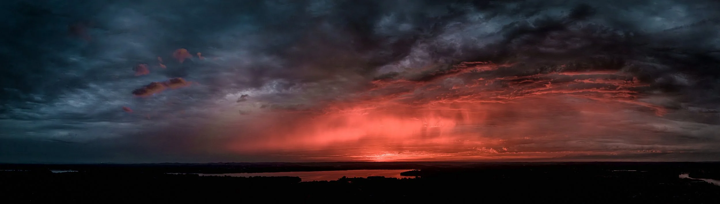 A dramatic sunset over a landscape with dark clouds, casting a red glow across the sky and reflecting on the water below.