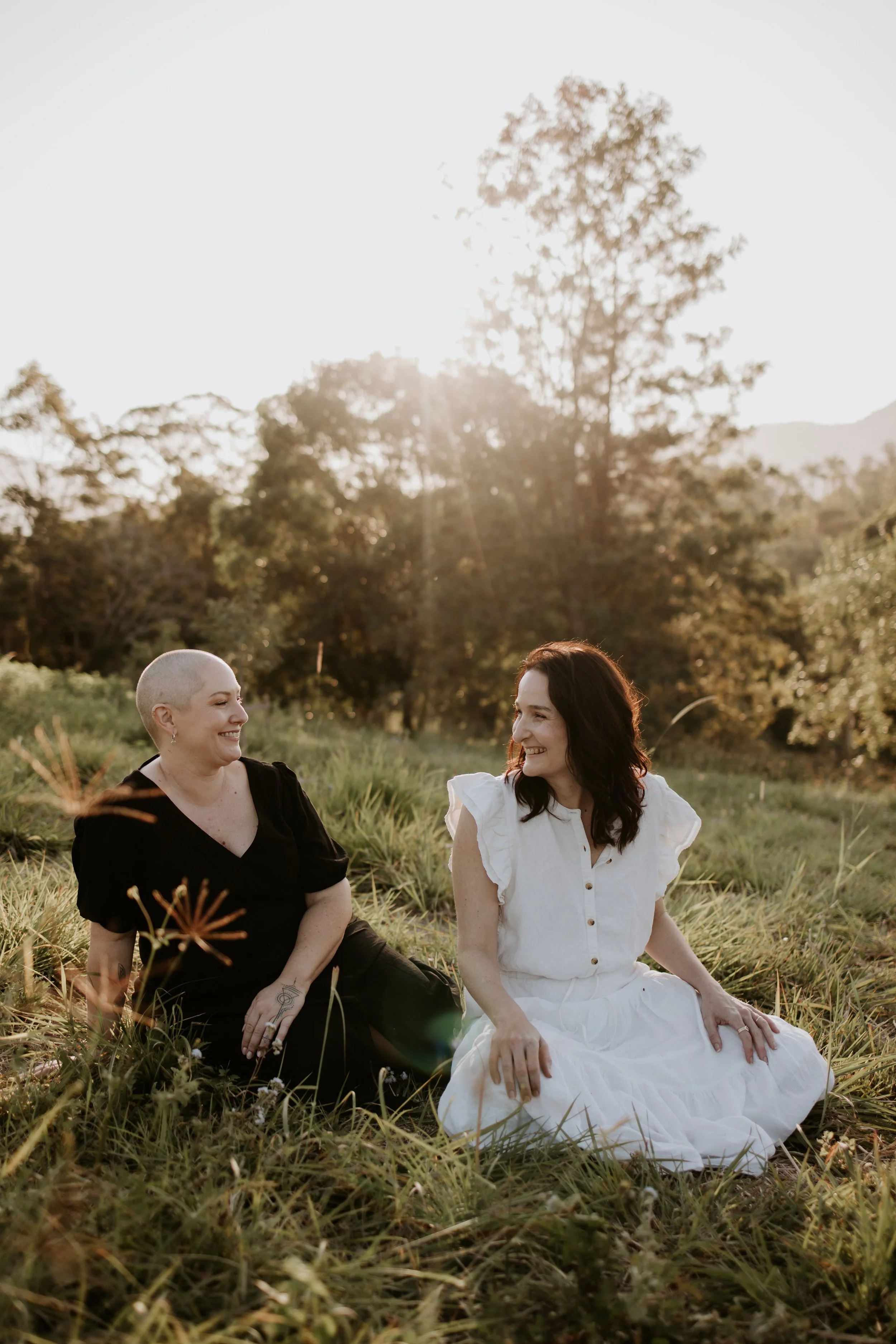 Two women sitting on grass in a field during sunset, smiling and looking at each other, with trees and hills in the background.