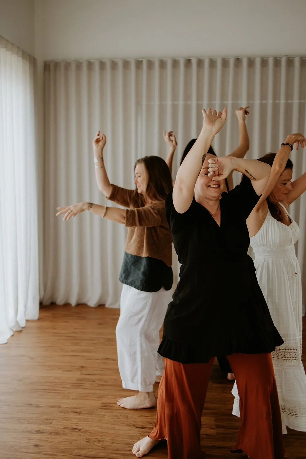 Women dancing indoors with white curtains in the background and wooden floor.