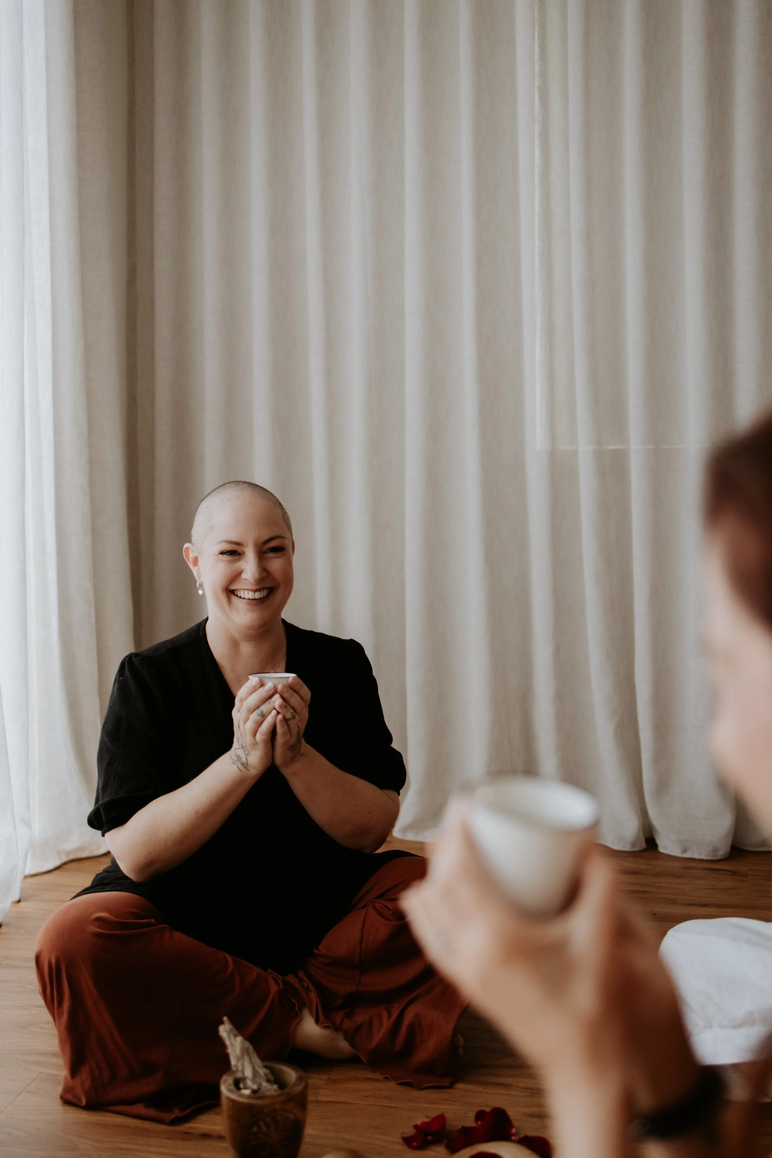 A woman with a shaved head and earrings smiling and holding a white cup, sitting cross-legged on a wooden floor with a candle, rose petals, and another person holding a cup in front of her, in front of cream-colored curtains.