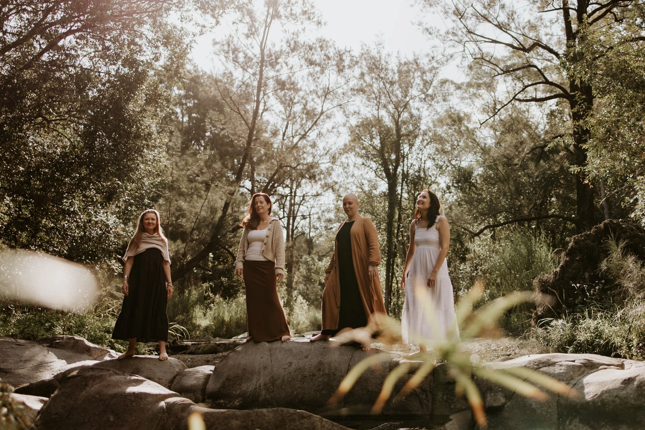 Four women standing on rocks near a creek in a forest, with sunlight filtering through trees.