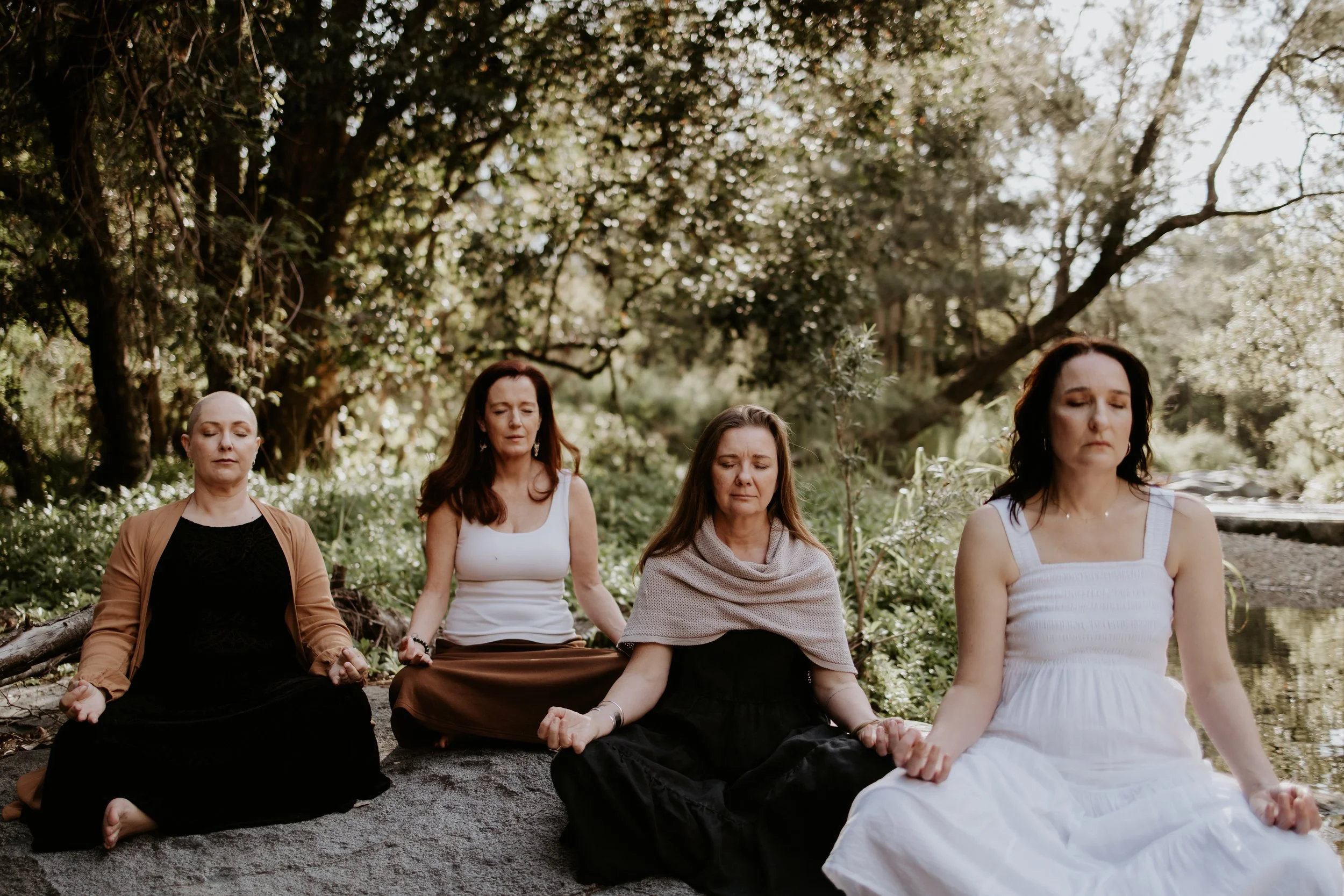 Five women sitting cross-legged outdoors in meditation with closed eyes, surrounded by trees and greenery.