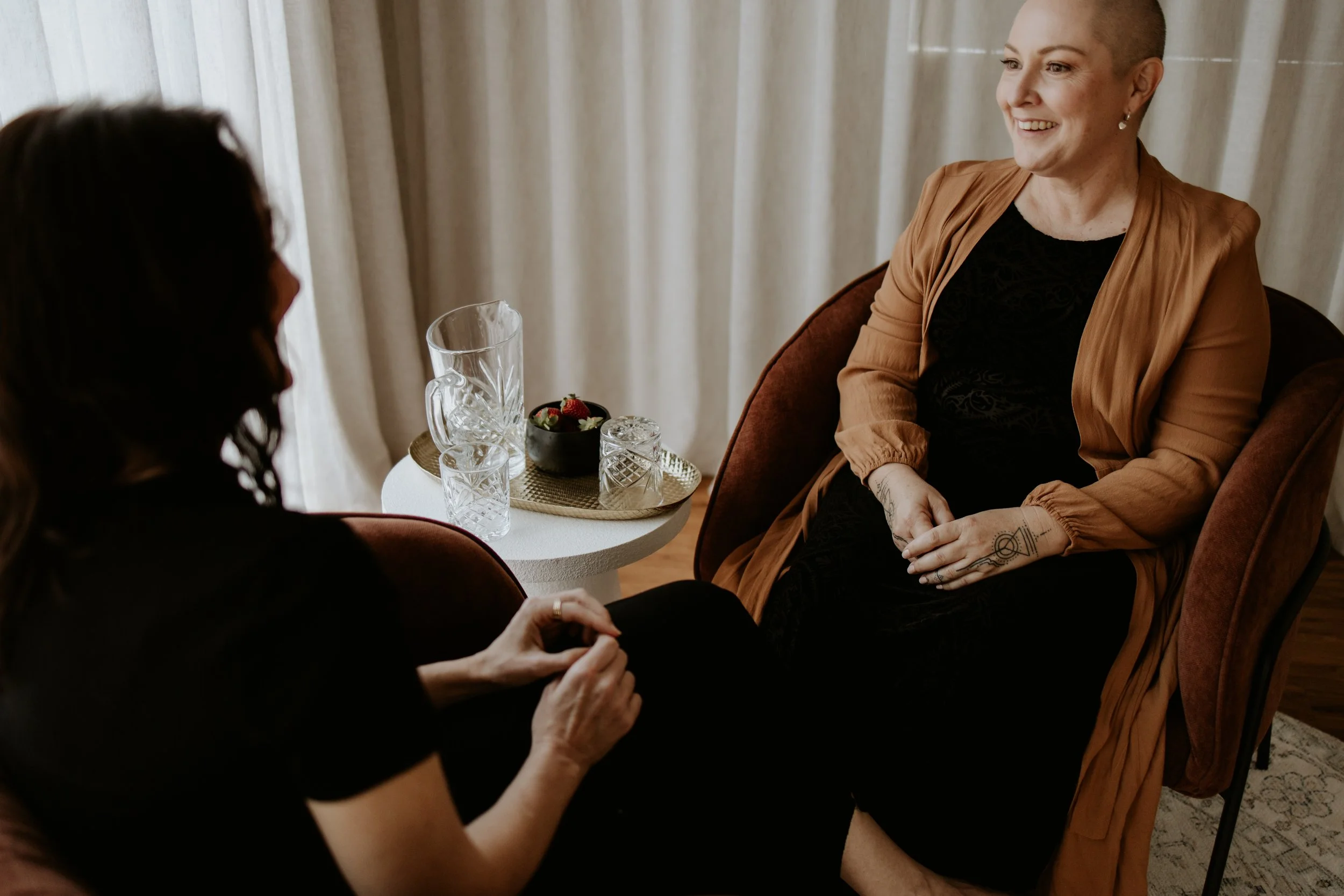 Two women sitting and talking in a cozy room, one with a shaved head and tattoos wearing a brown jacket, the other with long dark hair, and a small round table with glassware and strawberries between them.