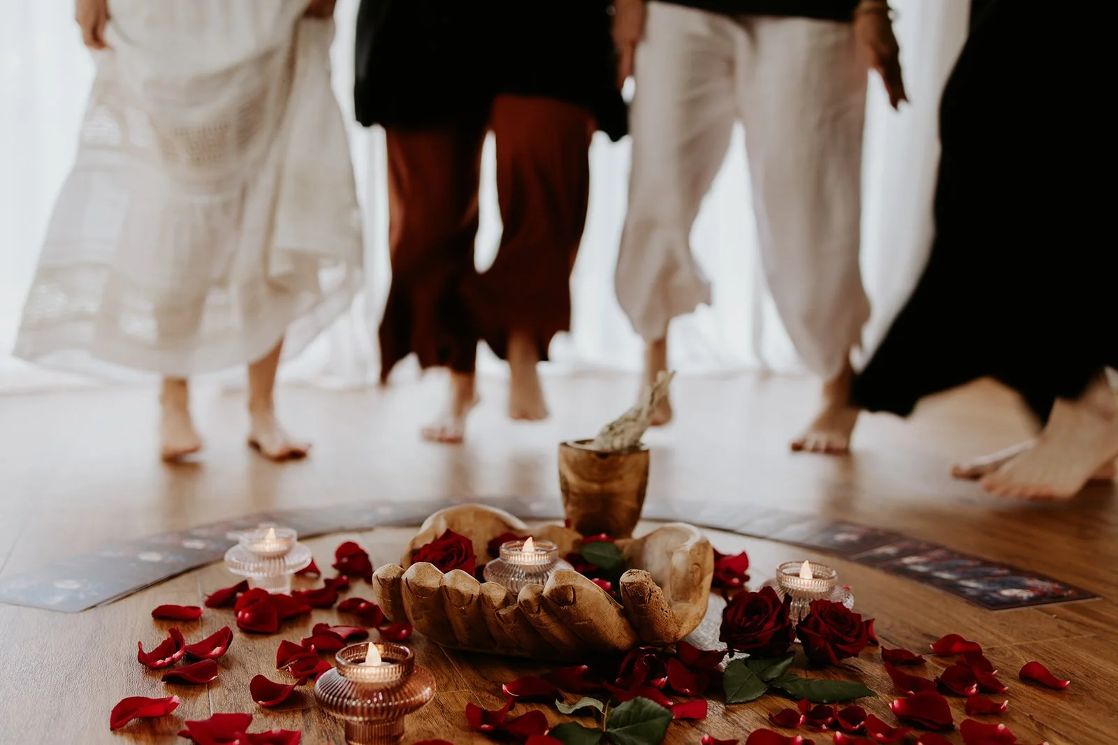 People walk barefoot around a ceremonial altar with candles, roses, stones, and a wooden cup on a wooden floor, possibly in a spiritual or ritual setting.