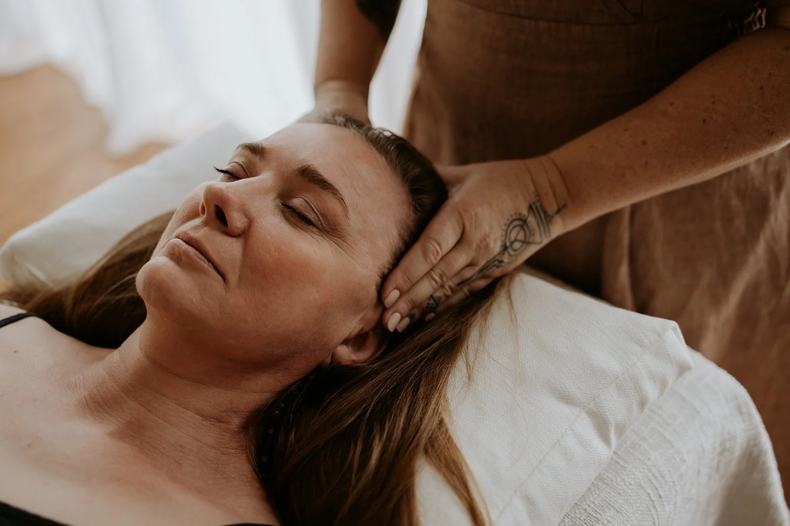A woman lying on her back with eyes closed, receiving a massage on her neck and head from a person with tattooed arms.