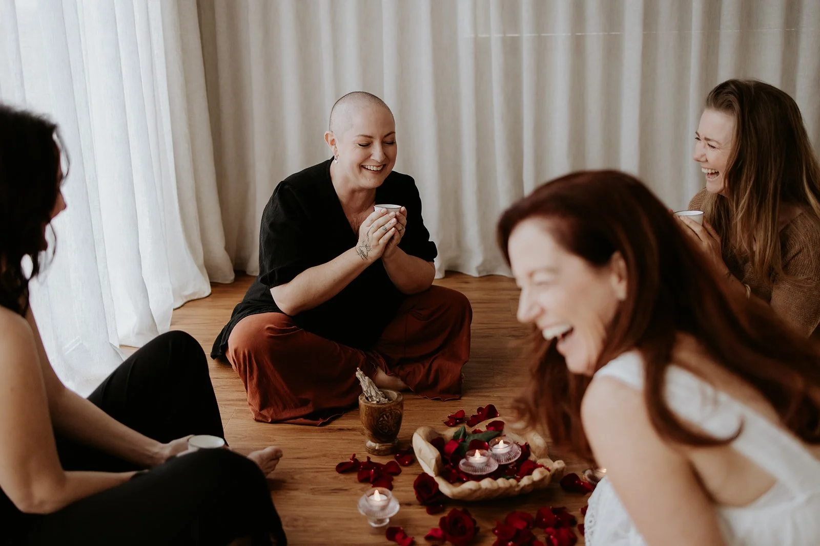 Group of women sitting on the floor in a circle, smiling and laughing, with candles, flowers, and a wooden bowl in the center, participating in a relaxing gathering or ceremony.