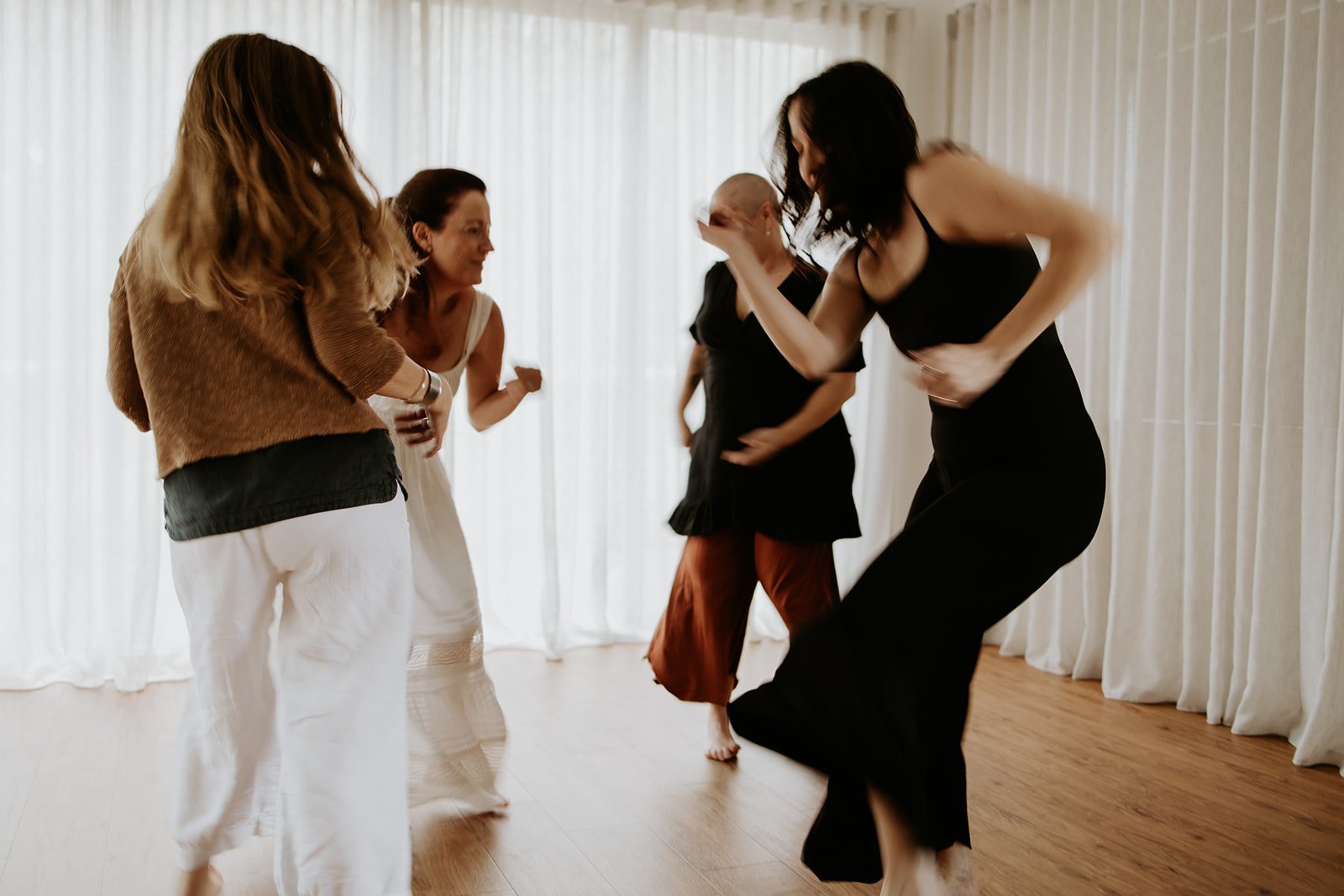 Four women dancing together in a room with light-colored curtains.
