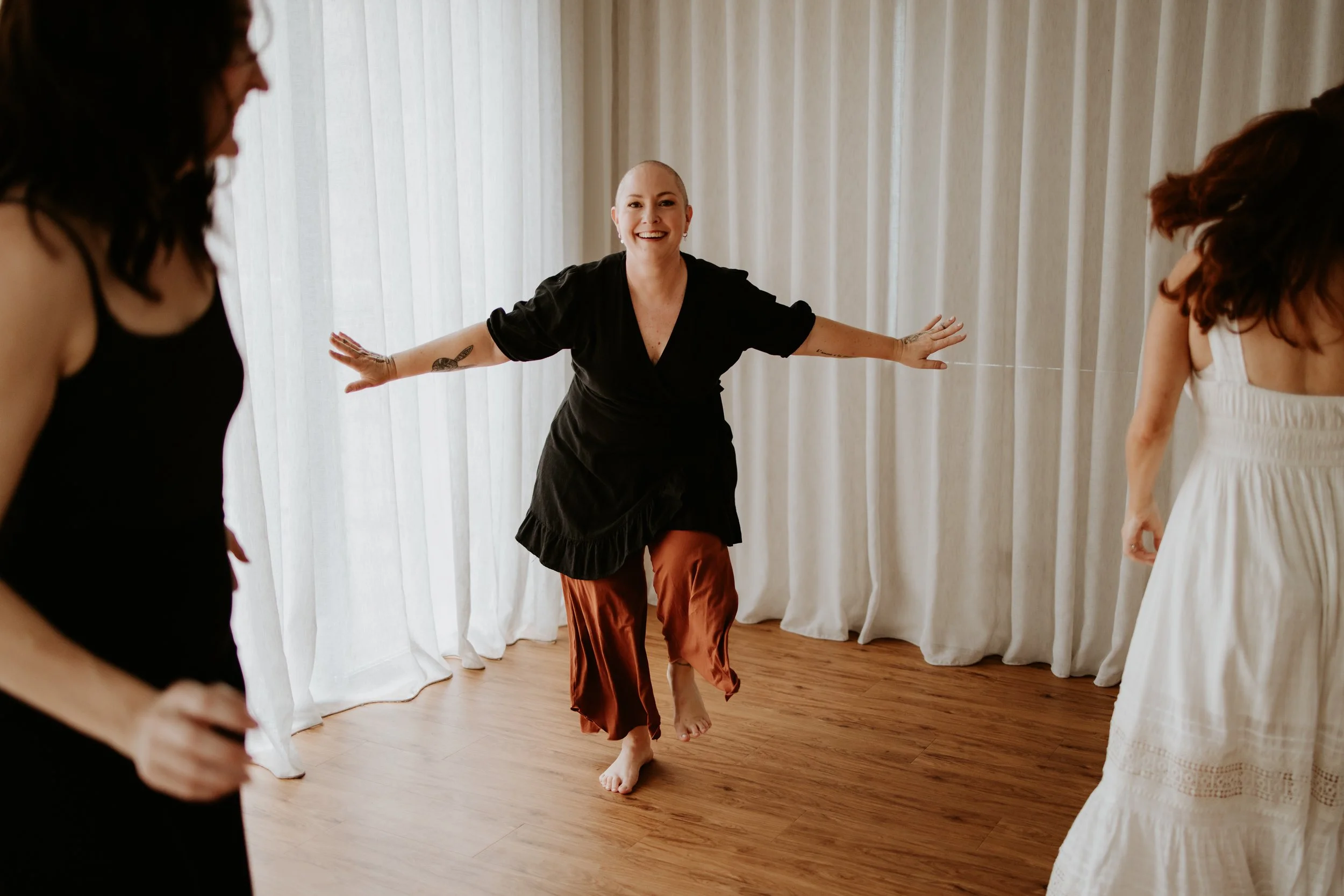 A woman with a shaved head, wearing a black dress and brown pants, smiling and dancing among other women in a room with white curtains and wooden floor.
