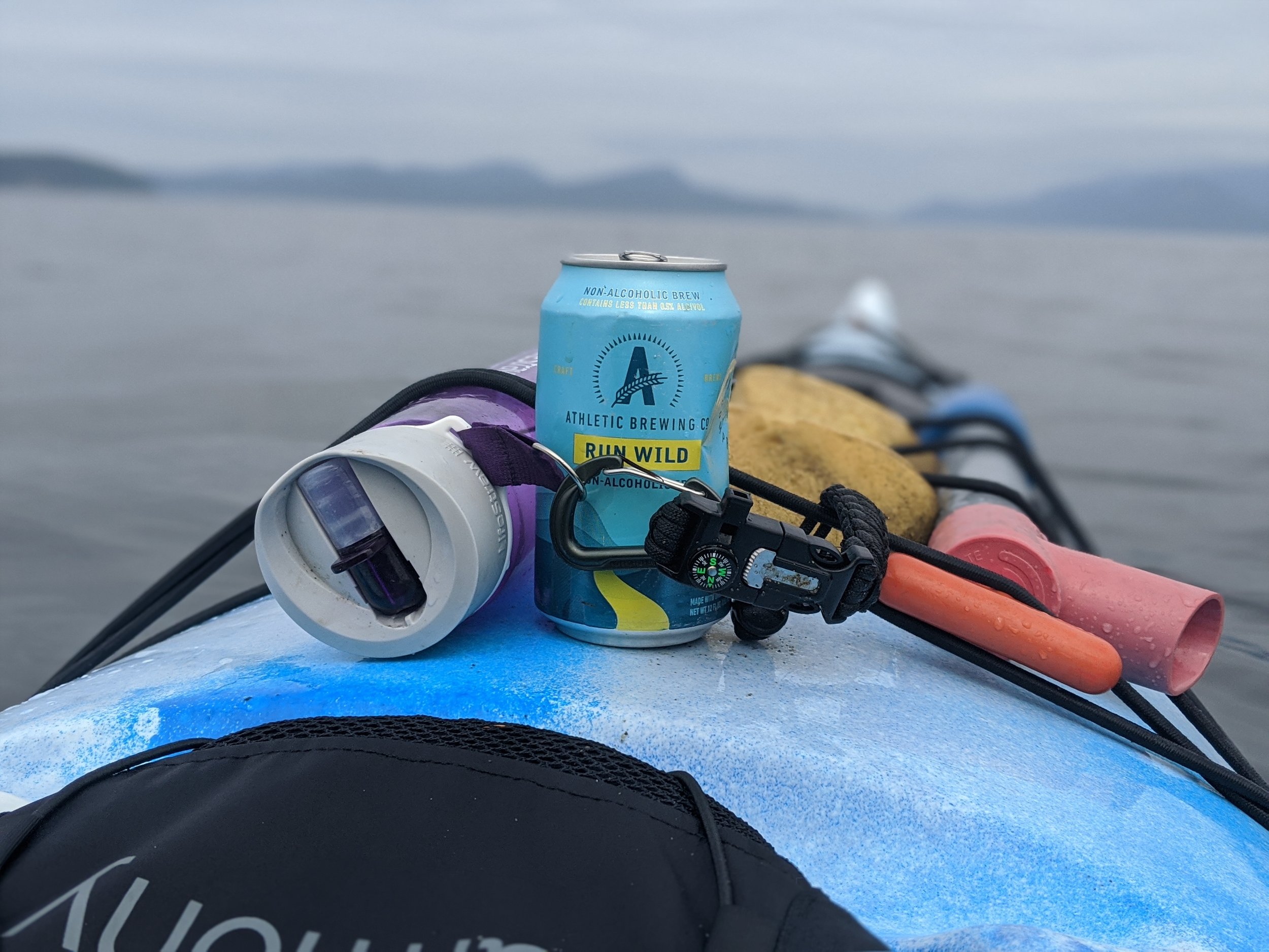 A close-up of a kayak deck with a can of athletic brewing beer, a black lanyard with a compass, a rolled-up sponge, and a purple water bottle top, floating on a body of water.