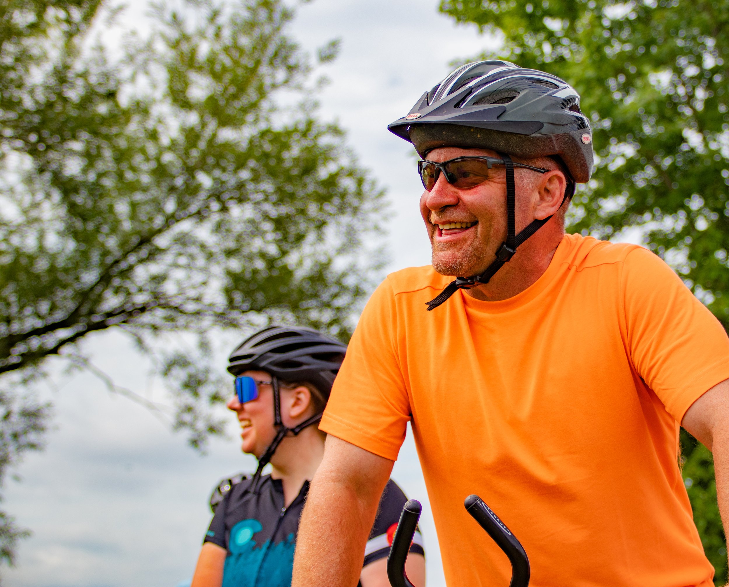 Two cyclists wearing helmets and sunglasses smiling outdoors, surrounded by trees.