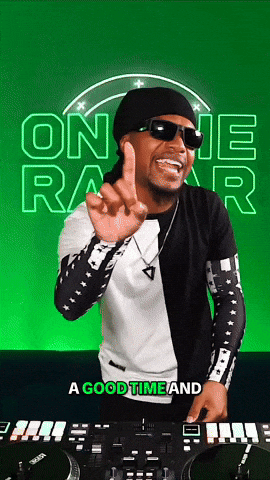 A smiling DJ wearing sunglasses and a black cap, pointing with his right hand, standing in front of a green neon sign that says 'ON THE RADER,' with DJ equipment in front of him.