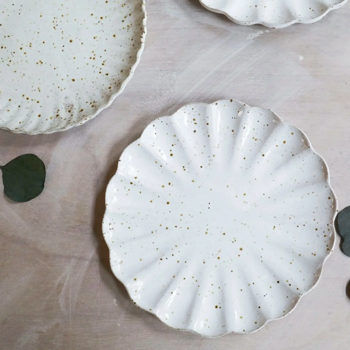 Three white ceramic plates with gold speckles and scalloped edges are placed on a light-colored wooden surface. Green leafy eucalyptus branches are also present on the surface.