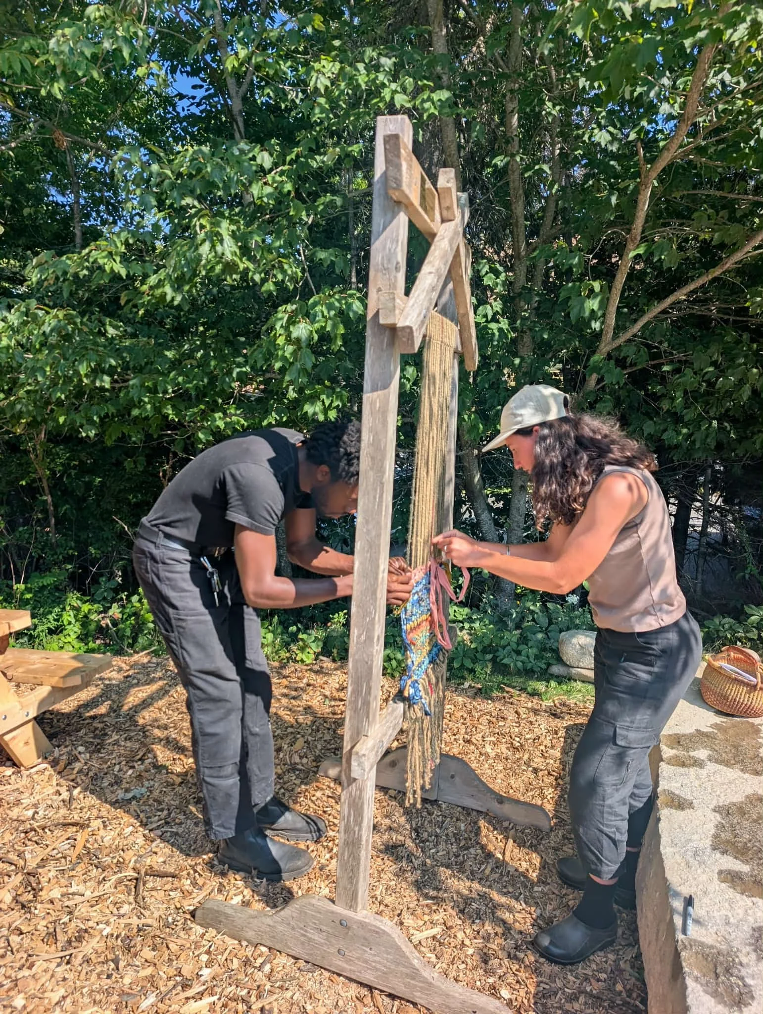 Two women are tying a colorful cloth on a wooden cross outdoors, with trees and green foliage in the background, on a sunny day.