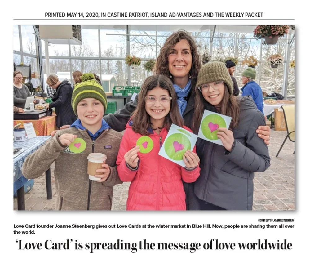 Four people, including an adult woman and three children, smiling and showing love cards with pink hearts on them inside a greenhouse or garden center. There are tables with items in the background and other people shopping.