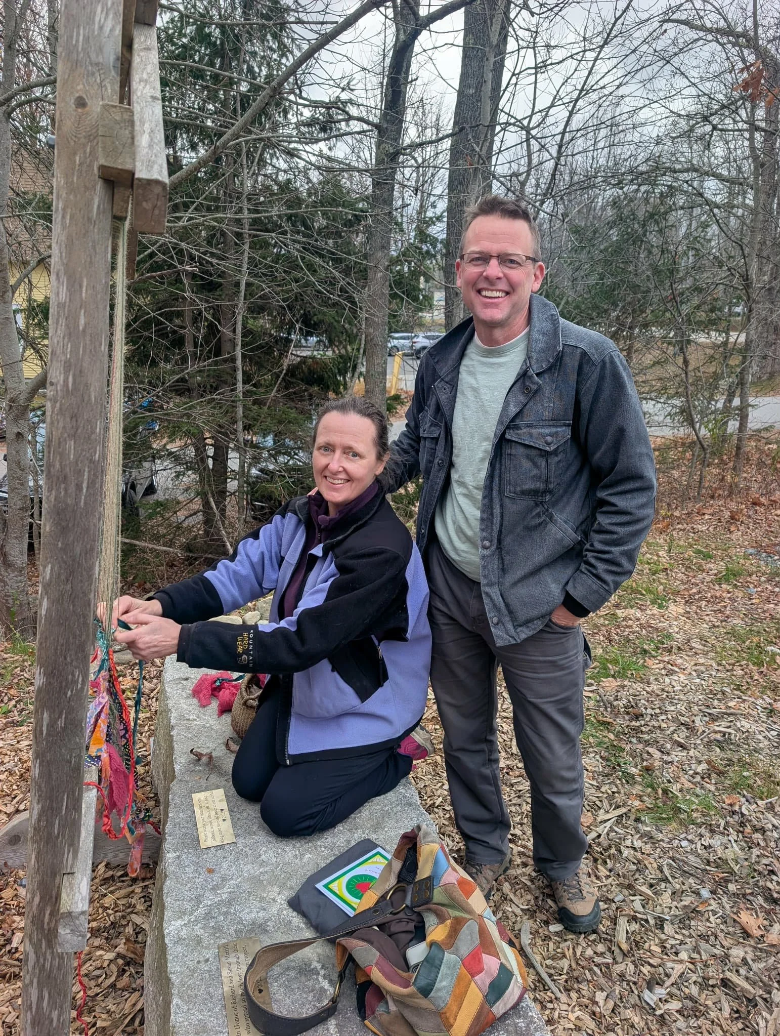 Two people, a woman kneeling and a man standing, smiling outdoors in a wooded area during fall, near a stone monument with a plaque and colorful strings tied to a wooden post.