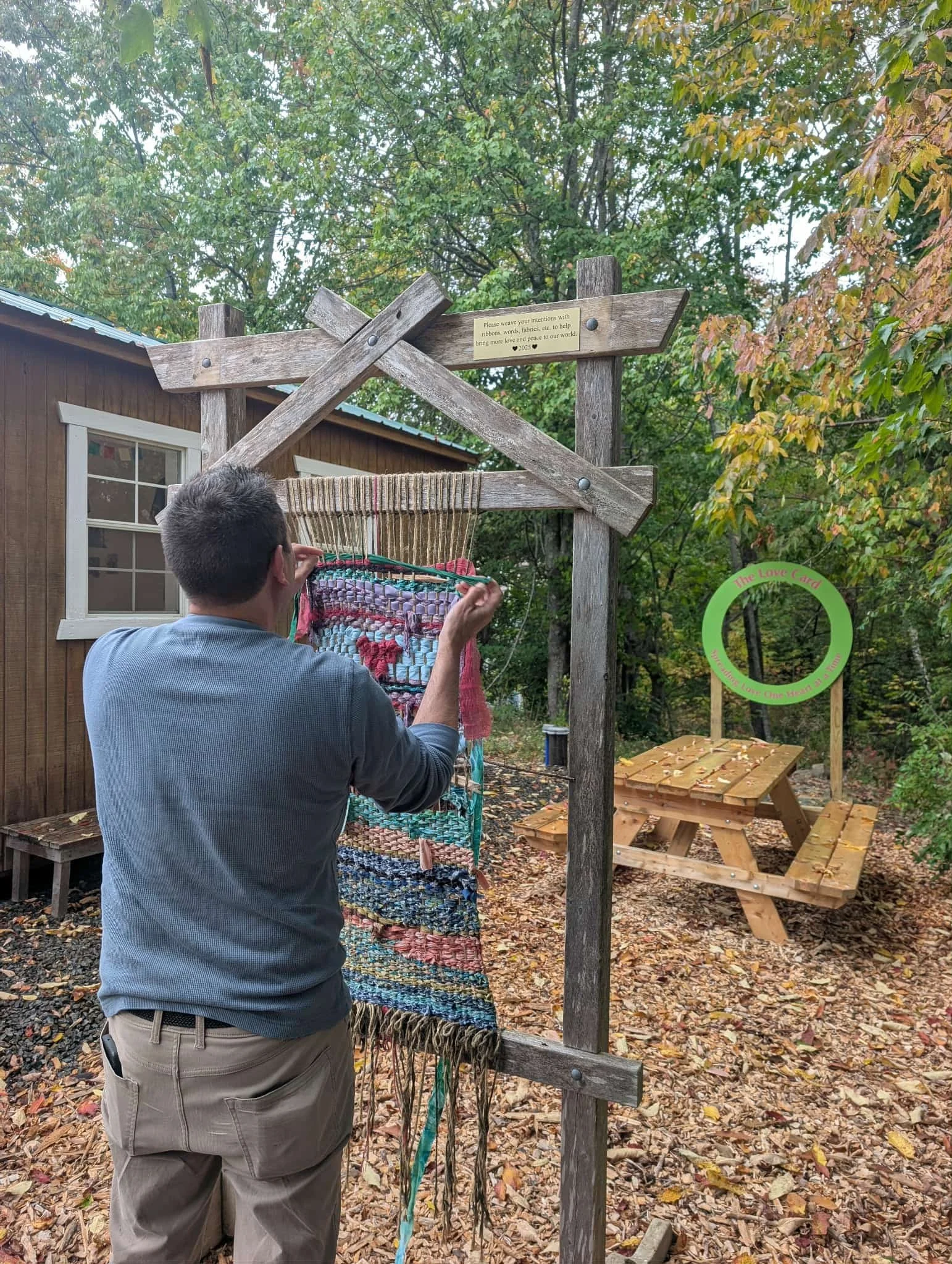 A man weaving a colorful textile on a wooden frame outdoors, surrounded by autumn trees and a small wooden building.