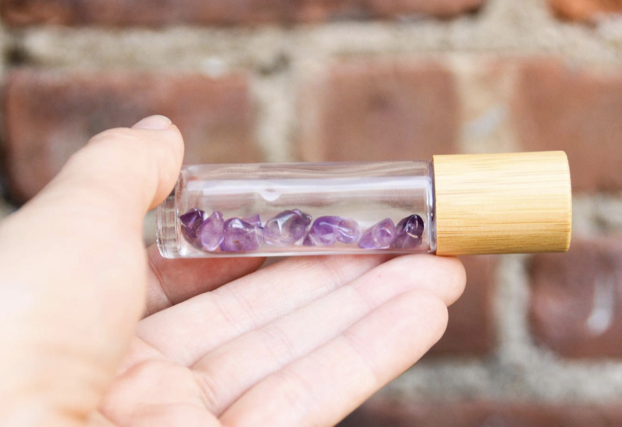 Hand holding a clear vial with small amethyst crystal chips inside and a wooden cap, brick background.