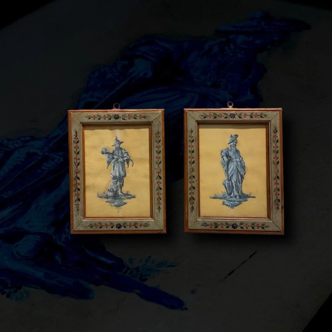 Two framed paintings of stylized blue and white statues of women, with floral borders, placed on a dark surface.