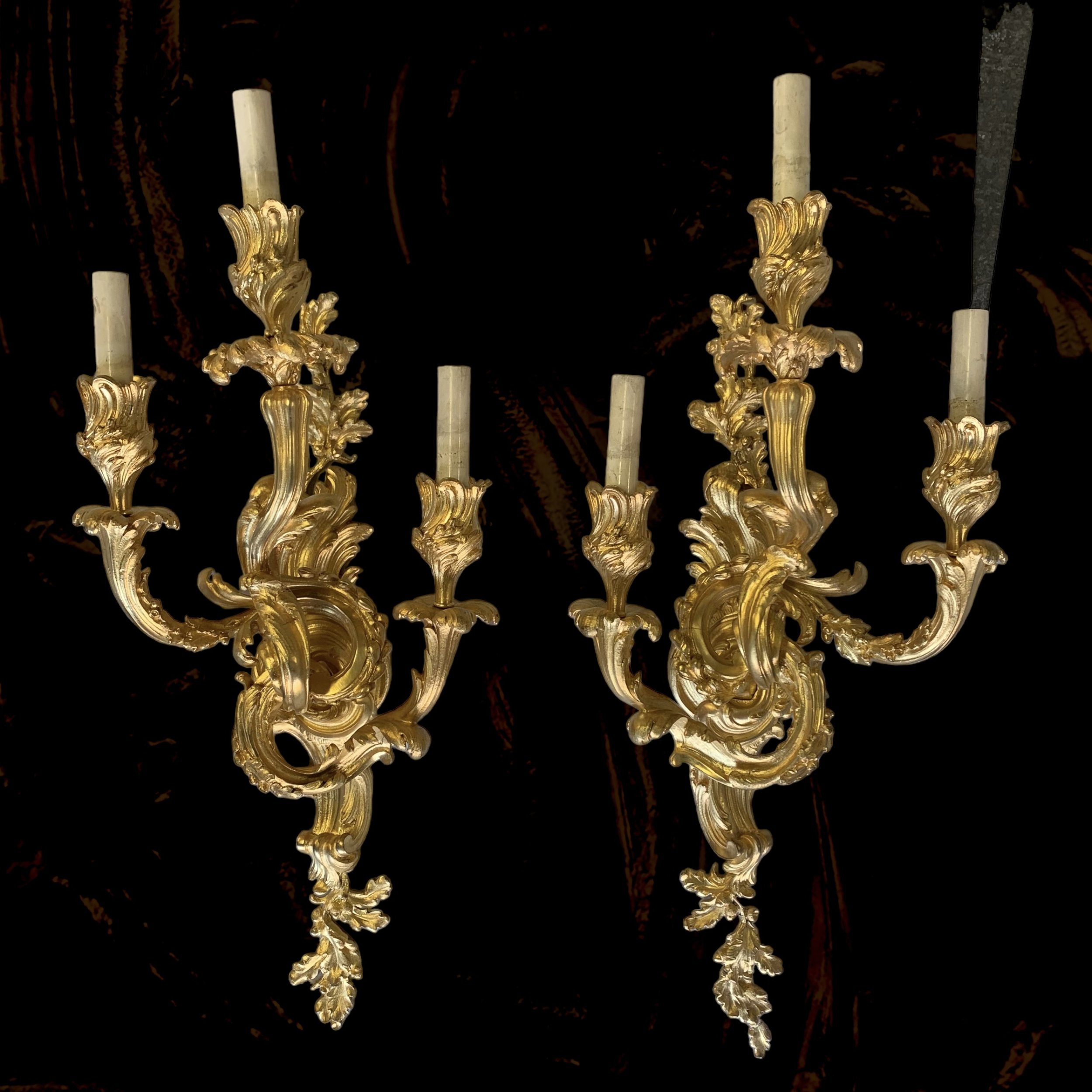 Gold ornate wall sconces with candleholders and white candles against a dark background.
