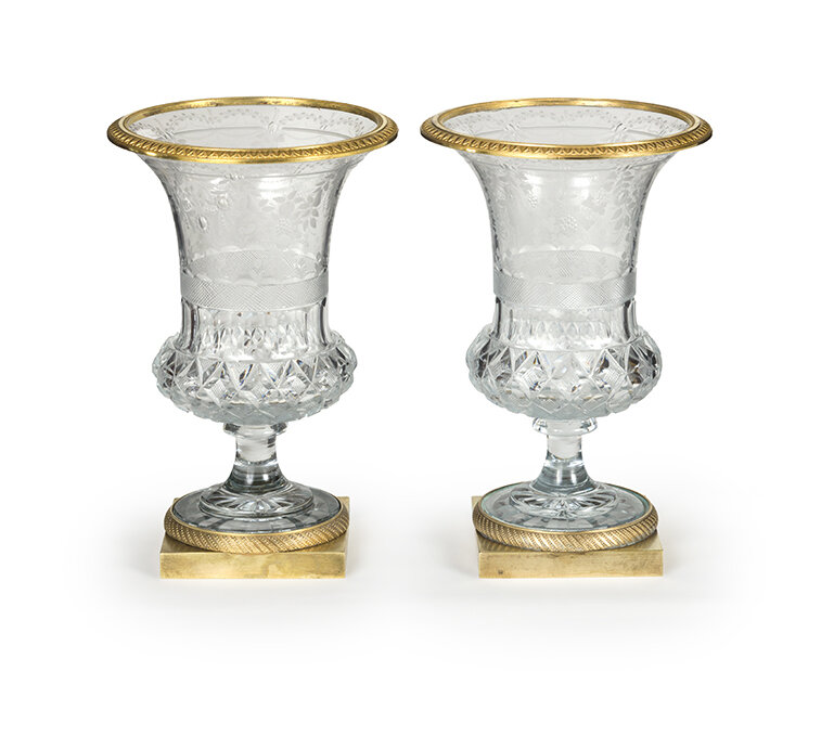 Two ornate glass goblets with gold trim on the rim and base, placed on small gold-colored square platforms.