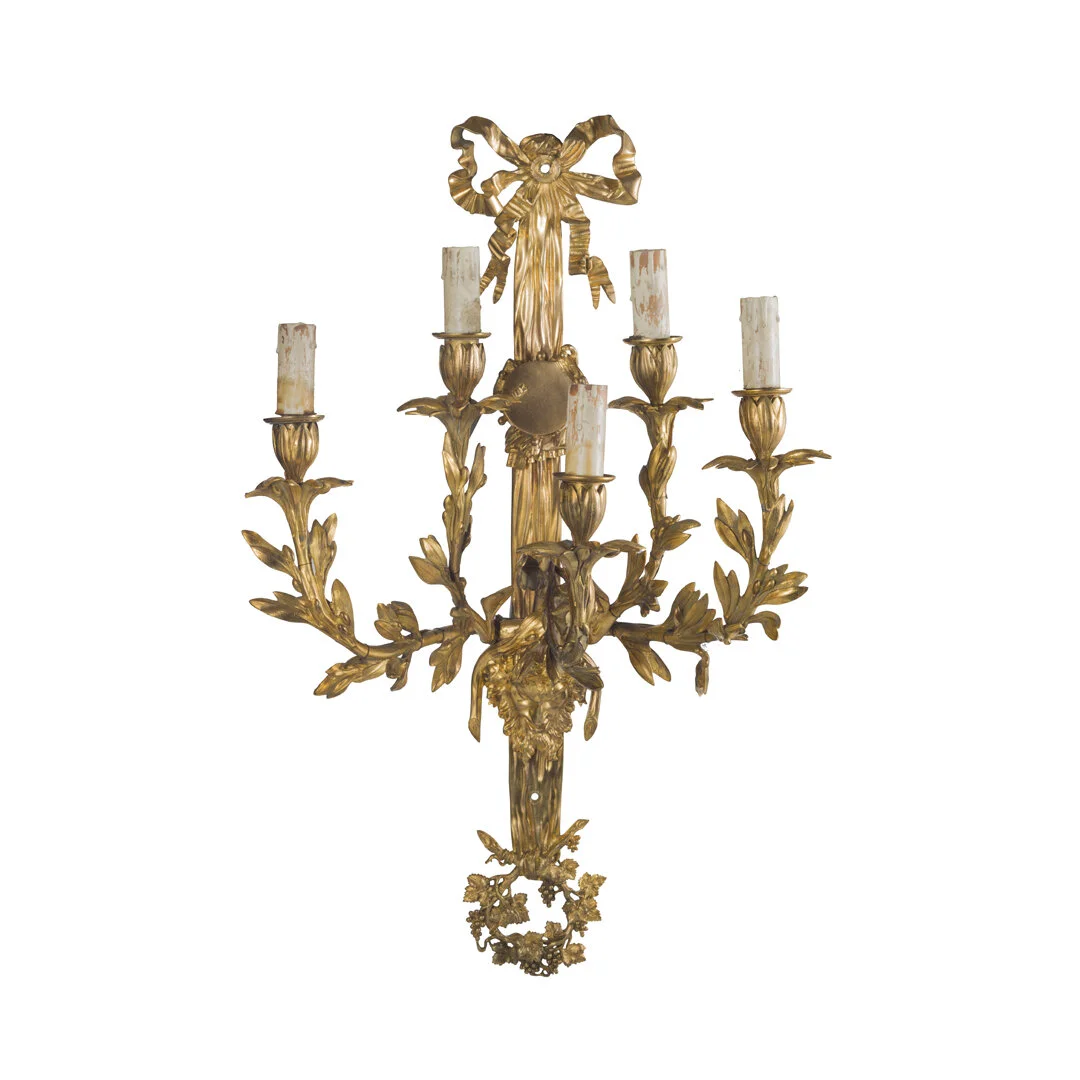 Gold-colored wall sconce with four candle holders, decorated with a bow and leaf motifs.