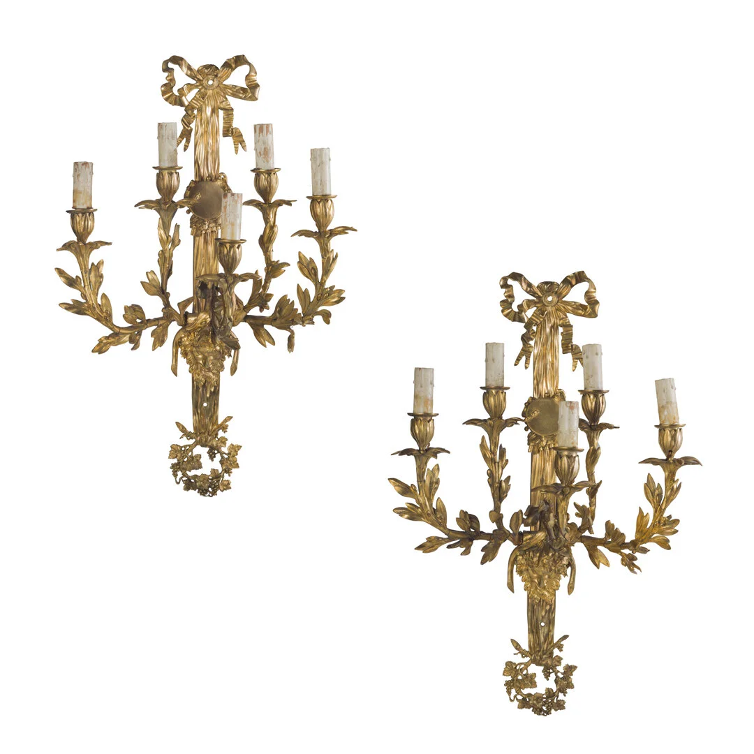 Two ornate gold wall sconces with three candle holders each, decorated with bows and leaf motifs, mounted on a white background.
