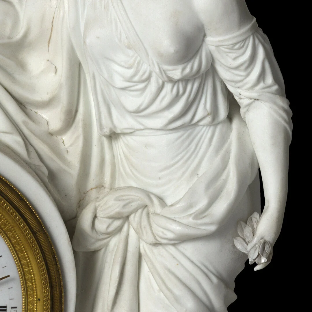 Close-up of a white ornate statue of a woman wearing a draped gown, holding a flower, with a partial view of a clock to the side.