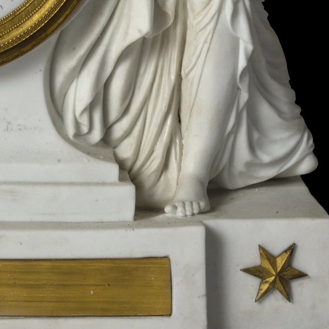 Close-up of a white marble sculpture of a person’s lower body and legs, with detailed drapery in their clothing, standing on a pedestal with a gold star decoration at the base.