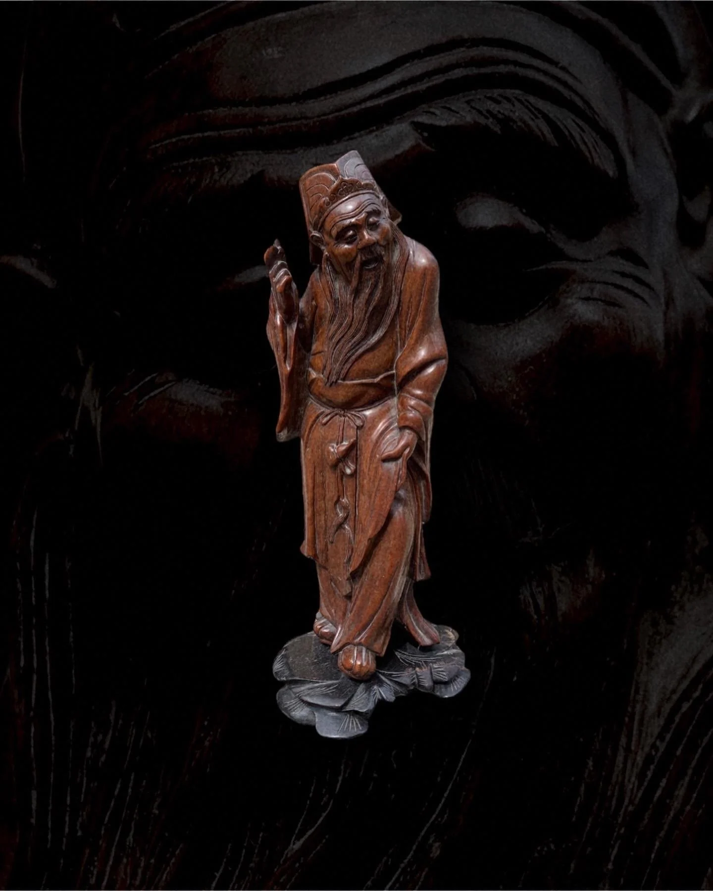 A detailed wooden sculpture of an elderly man with a long beard, wearing traditional robes, standing on a base with leaf-like details, set against a dark background.