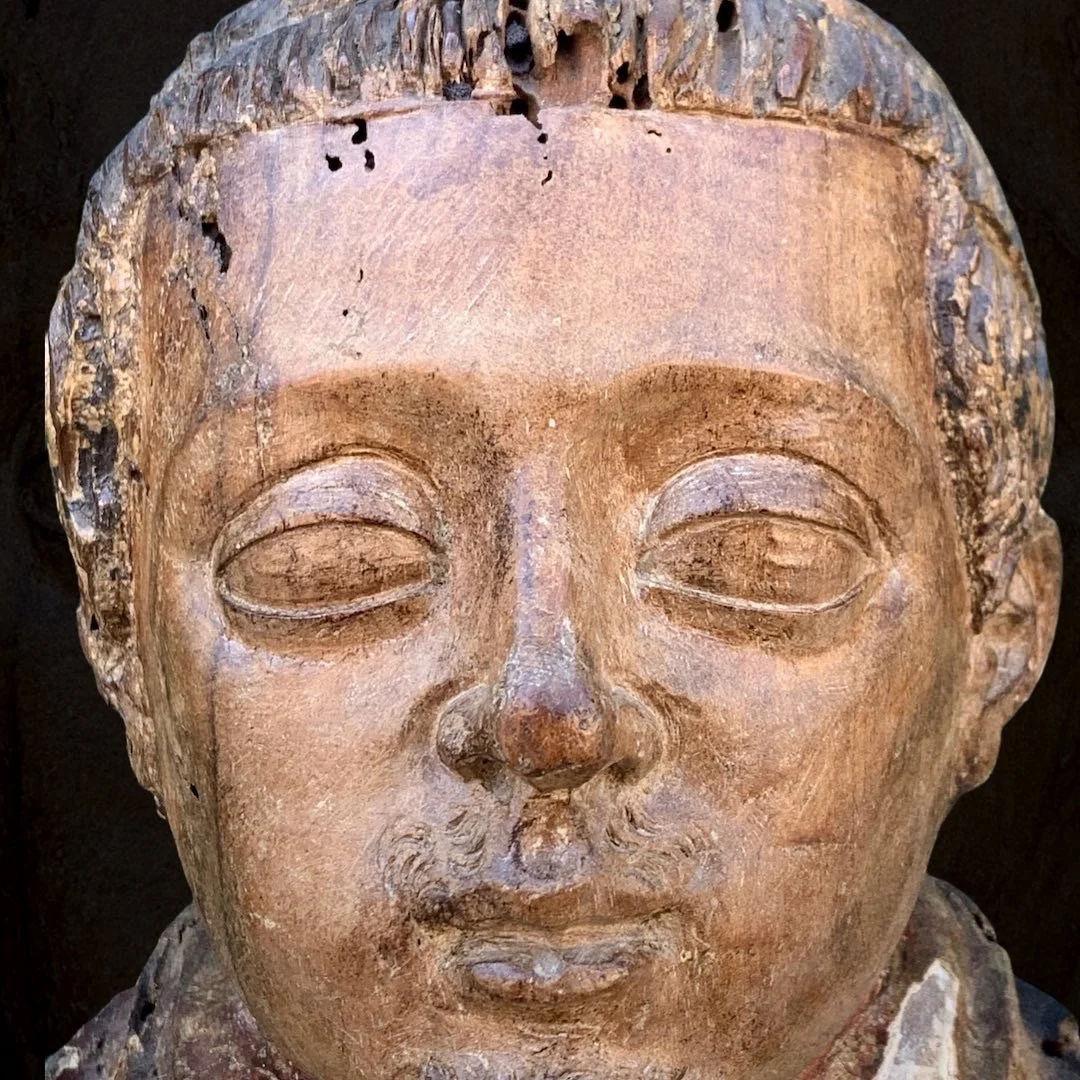 Close-up of an ancient wooden carved sculpture of a person's face with closed eyes, worn and weathered surface, some damage and small holes in the wood.