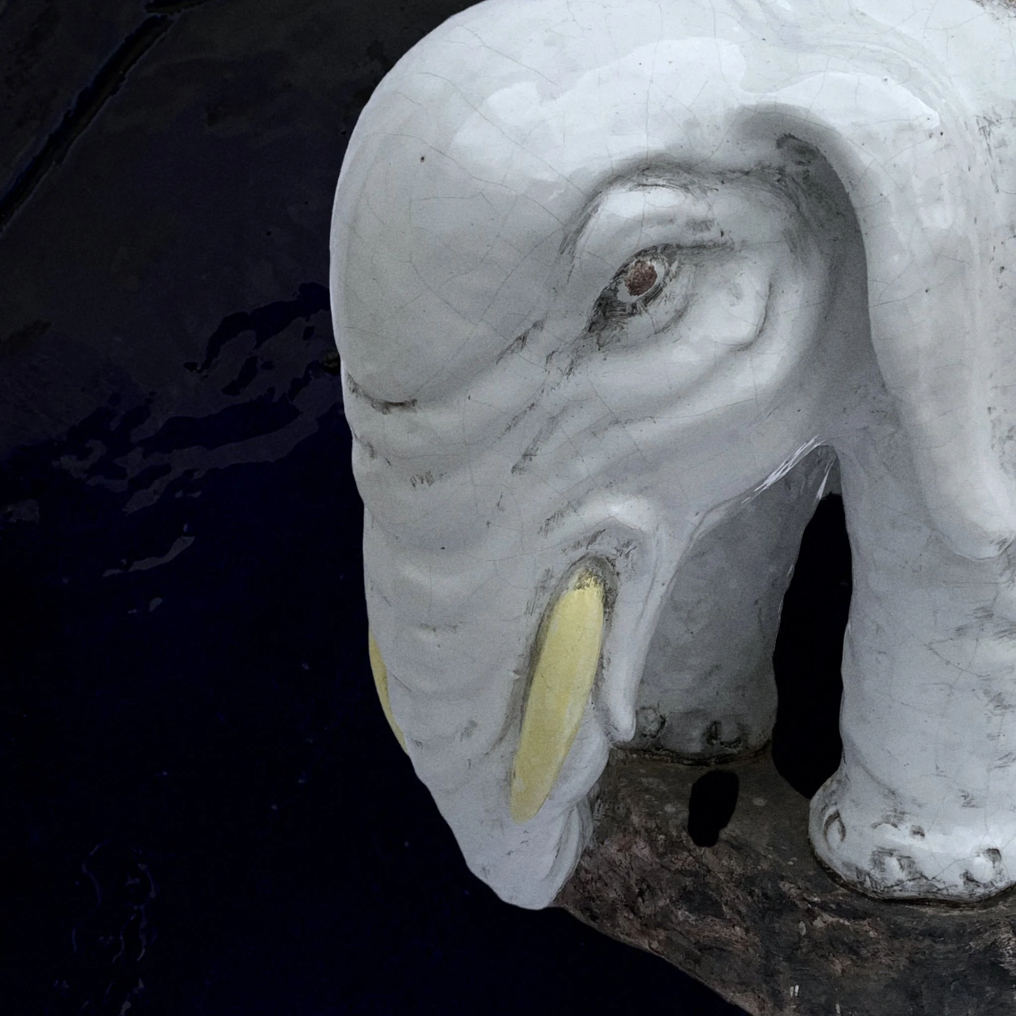 Close-up of a white carved wooden elephant head sculpture with yellow tusks, visible cracks, and a weathered surface, resting on a dark surface.