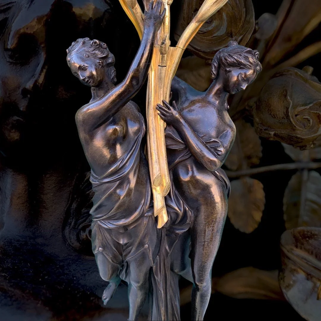 Close-up of a dark bronze sculpture of two women assisting each other, with intricate details on their dresses and hair, set against a background of large leaves.