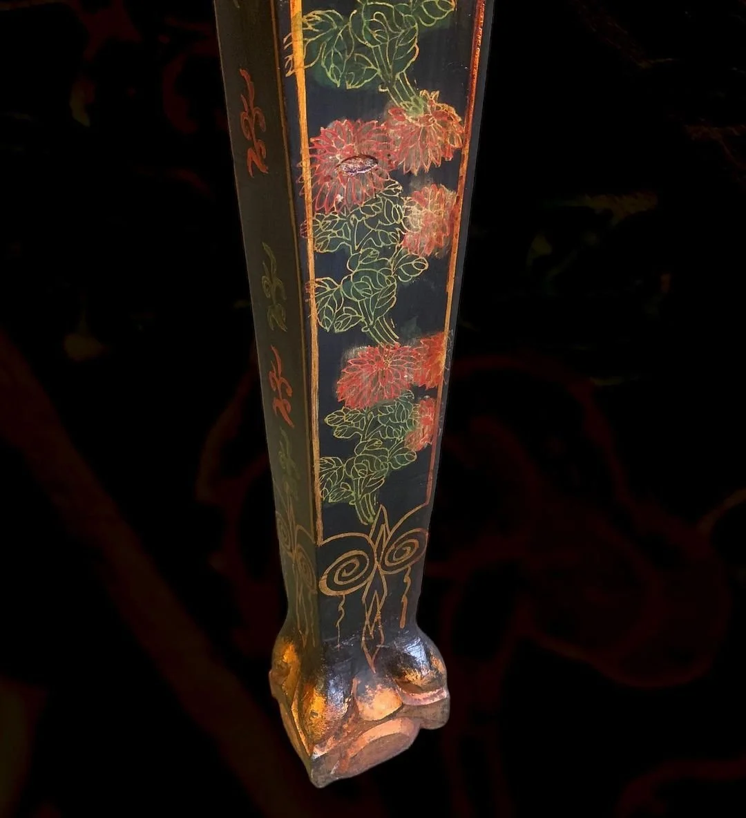 Close-up of a decorative, rectangular stand with floral and abstract designs painted in red, green, and orange on a black background, resting on a carved wooden base.