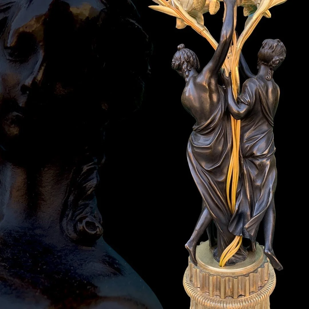 A detailed bronze sculpture of two women holding up a large plant or tree. The sculpture features intricate details of the women's dresses and hair, set against a black background.