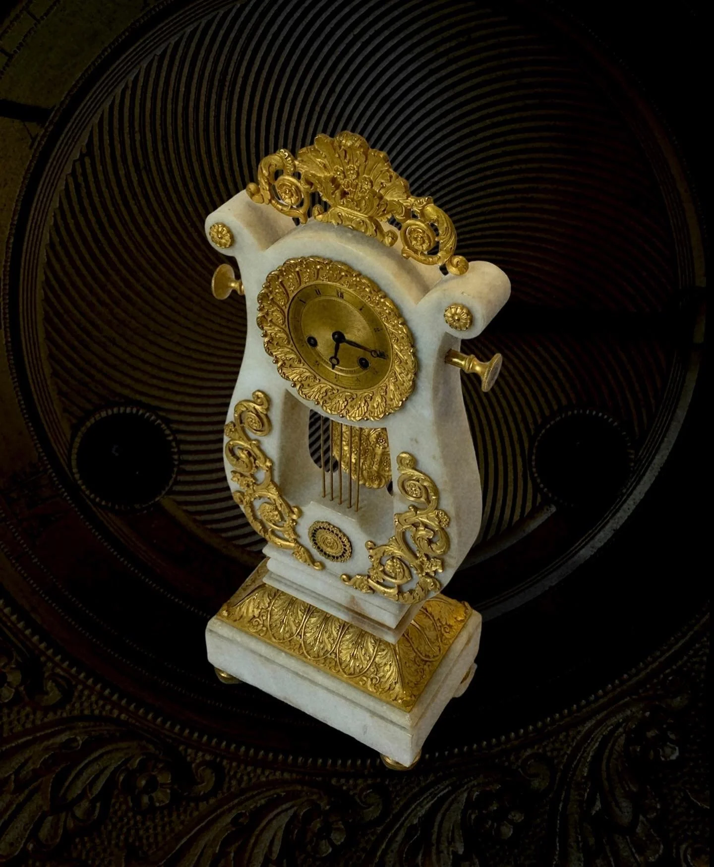 An ornate antique clock with gold decorations and a white marble body, placed on a decorative surface with a striped circular background.