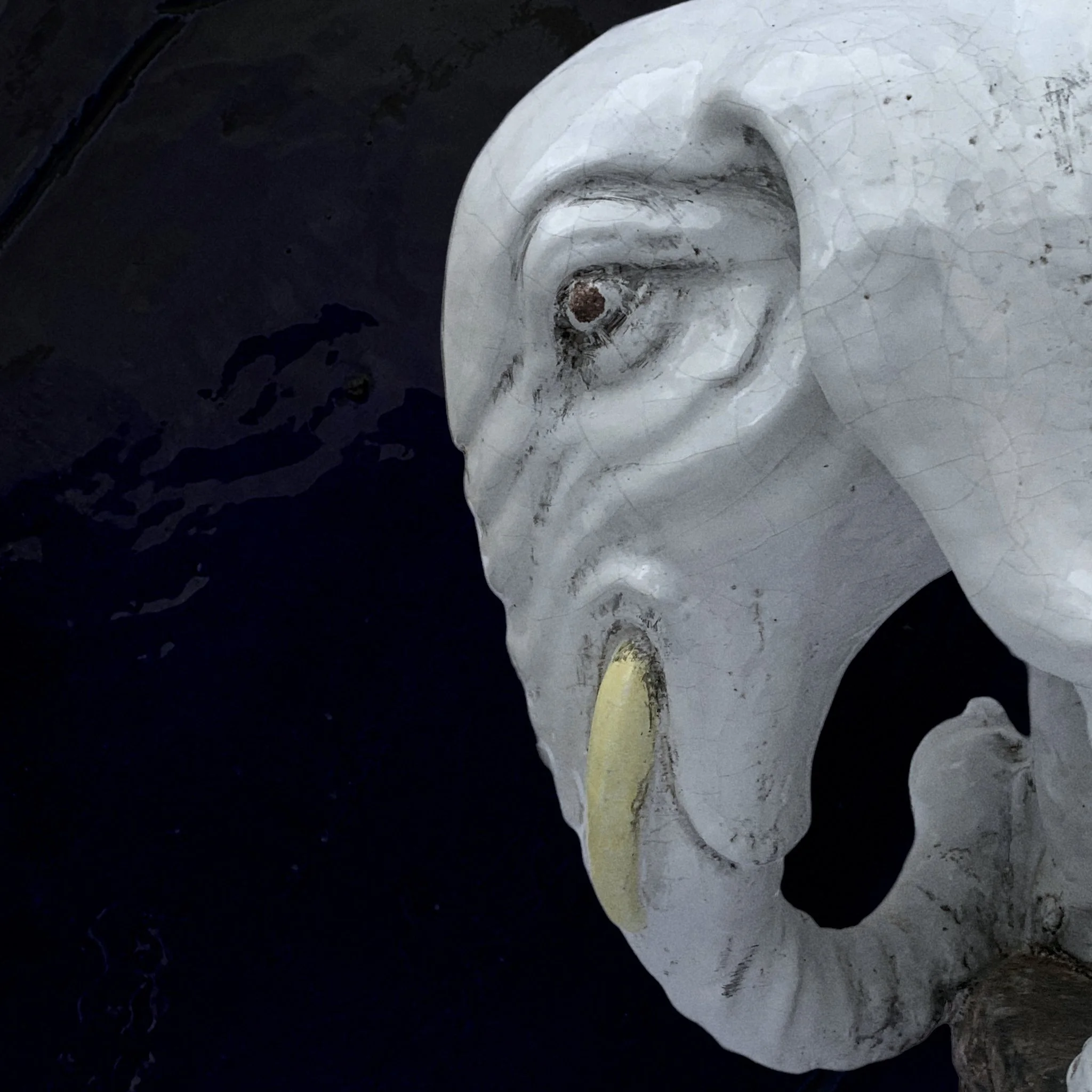 A close-up of a white, cracked stone sculpture of a face with weathered features, including a large nose, closed eyes, and an open mouth, with a dark background.