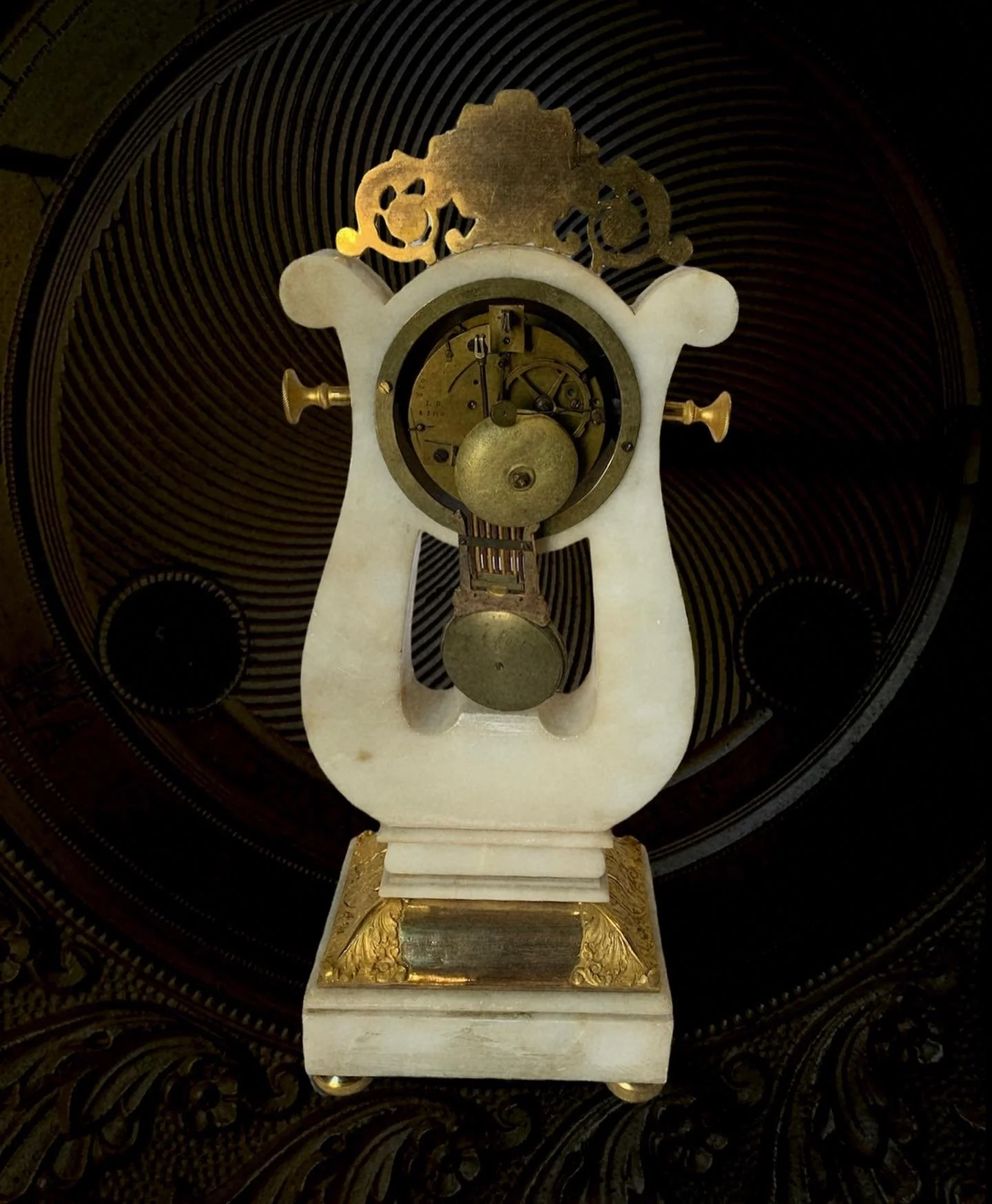 An antique clock with a white marble base and gold decorative details, with its internal gears and mechanisms visible, mounted on a patterned dark background.