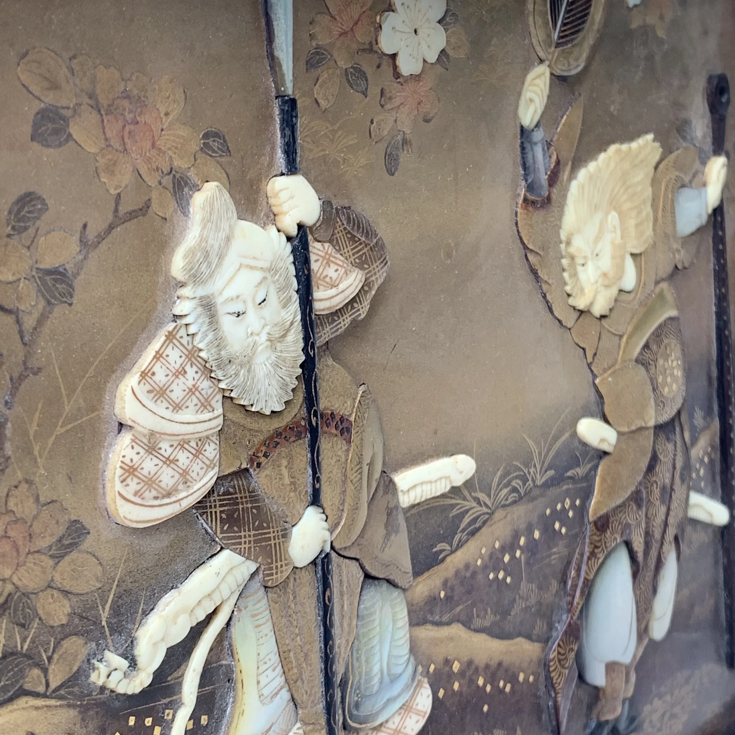 Close-up of a decorative item featuring carved ivory or bone figures, including a man with a beard and a hat holding a staff, with floral and landscape designs in the background.