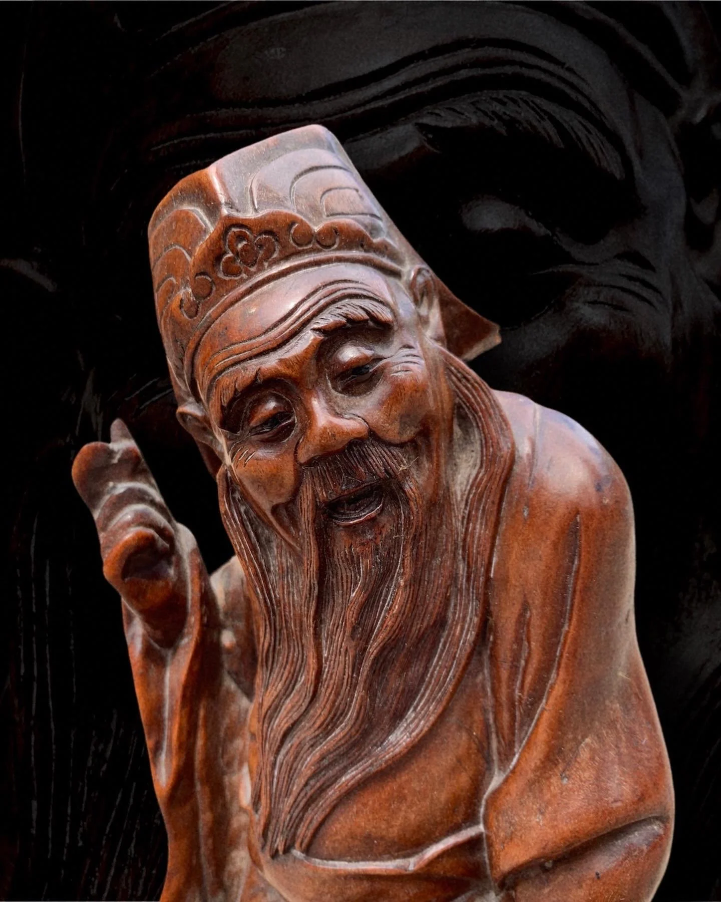 Wooden sculpture of an elderly man with a long beard, wearing a hat, with a joyful expression, against a dark background.