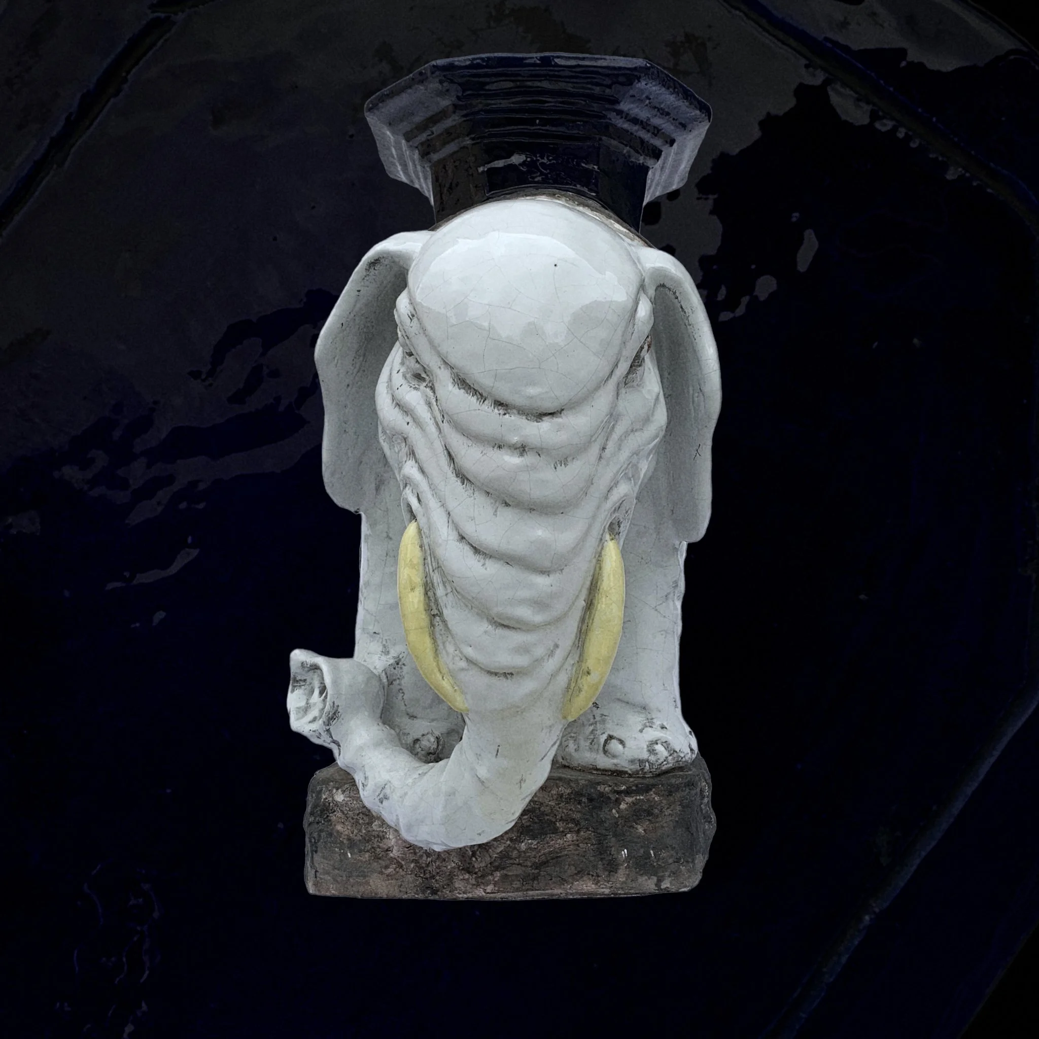 A white ceramic or plaster statue of an elephant with large yellow tusks, viewed from above, against a black background.