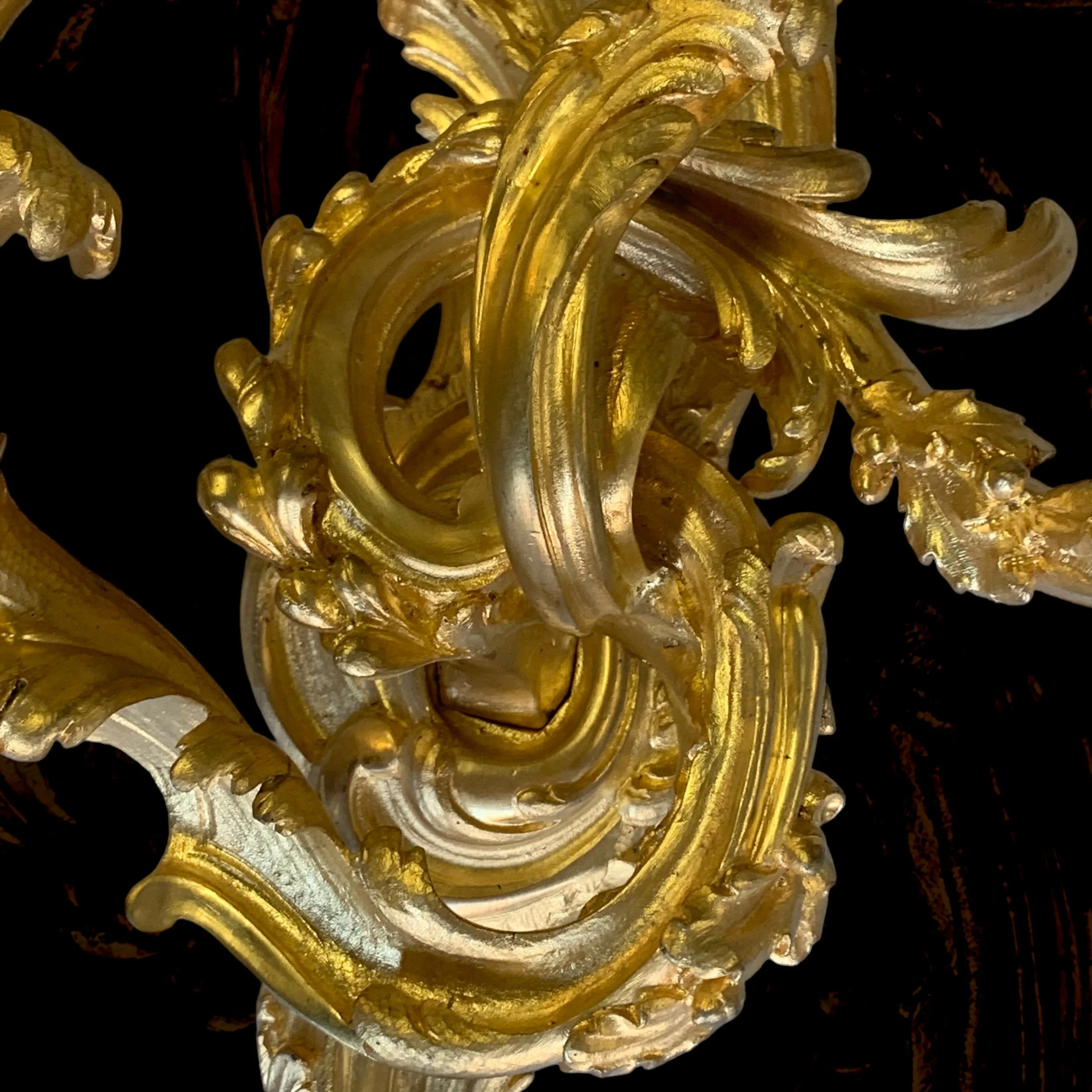 Close-up of an ornate, gold-colored decorative element with intricate scrollwork and leaf motifs against a dark background.