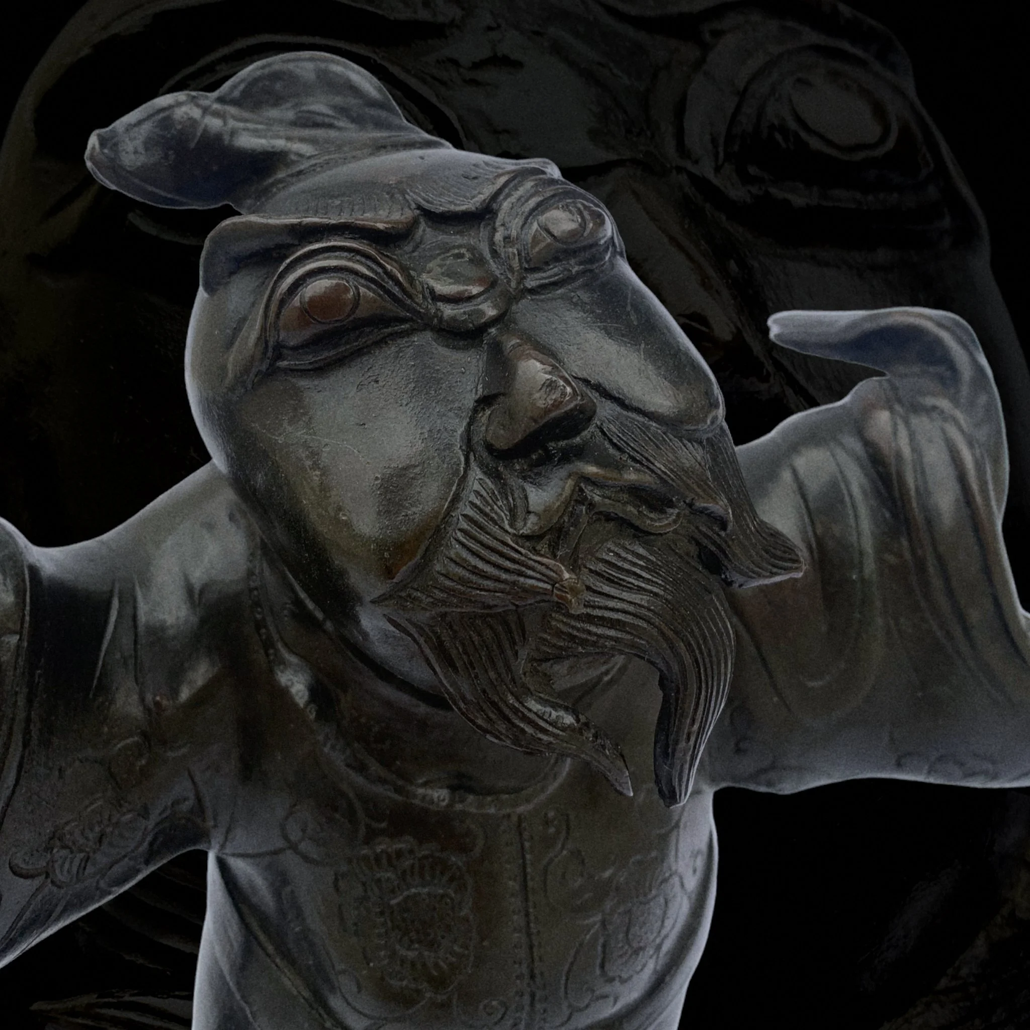 Close-up of a dark gray, carved sculpture of a bearded man with glasses, with outstretched arms, dark background.
